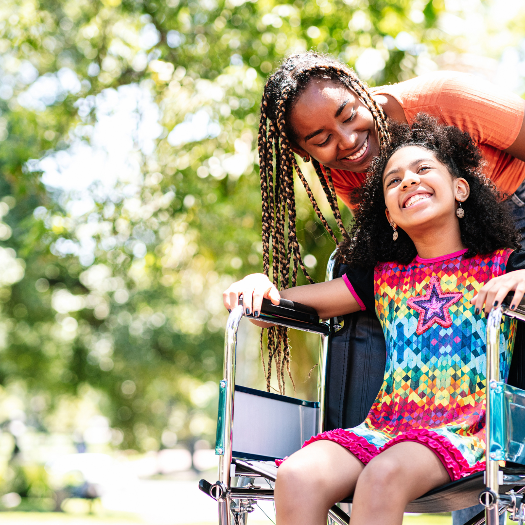 A young girl in a colorful dress smiling in a wheelchair outdoors, with a woman leaning over her affectionately.