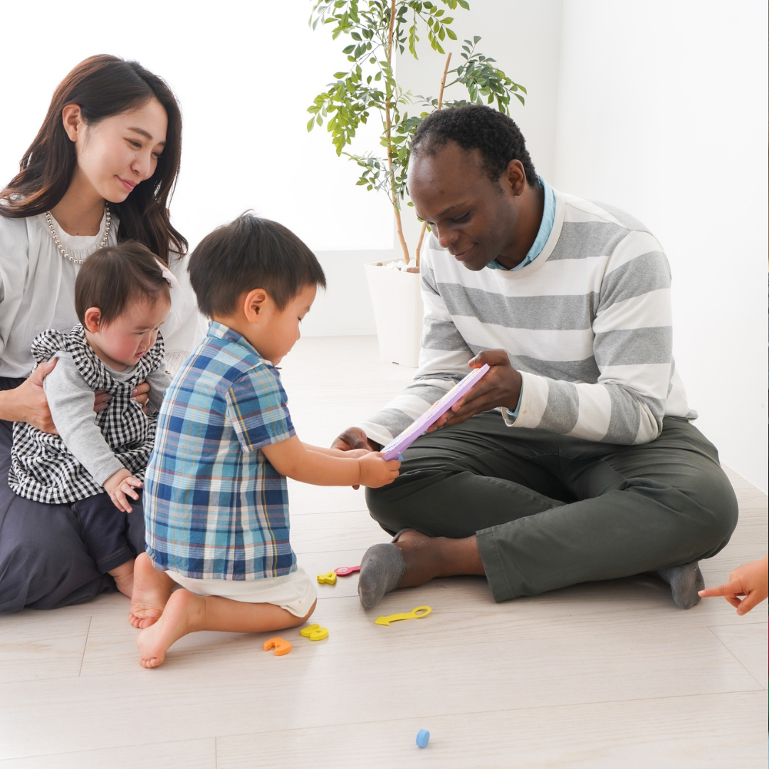 A diverse family spending quality time together indoors. The mother and children are sitting on the floor, engaging in a shared activity with a man, possibly their father. The children are focused on an object the man is holding, with toys scattered on the floor. A large potted plant is visible in the background.