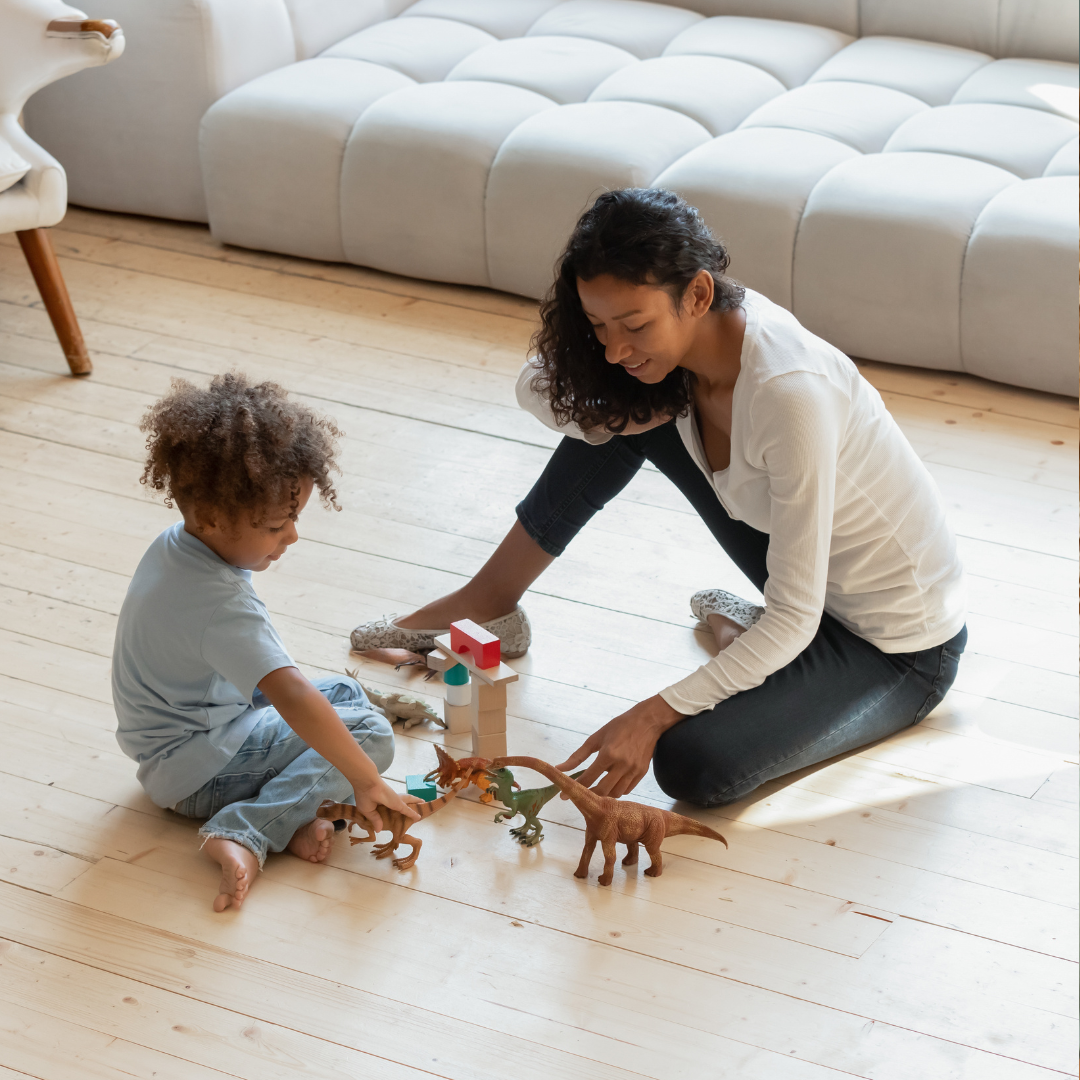 A woman and a young child playing with toy dinosaurs on a wooden floor in a living room.