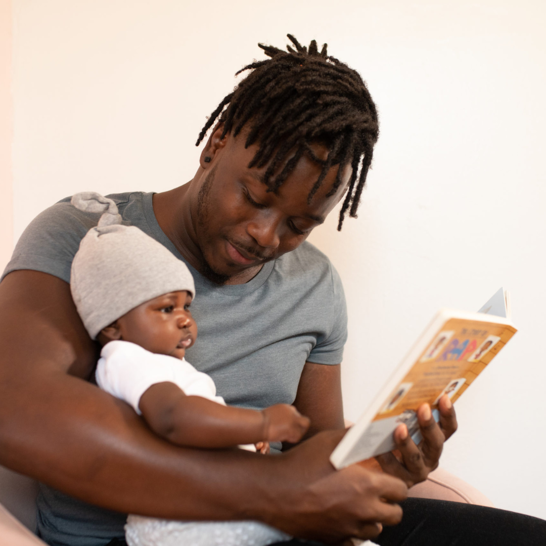 A man with dreadlocks holding a baby while sitting on a chair and reading a book together