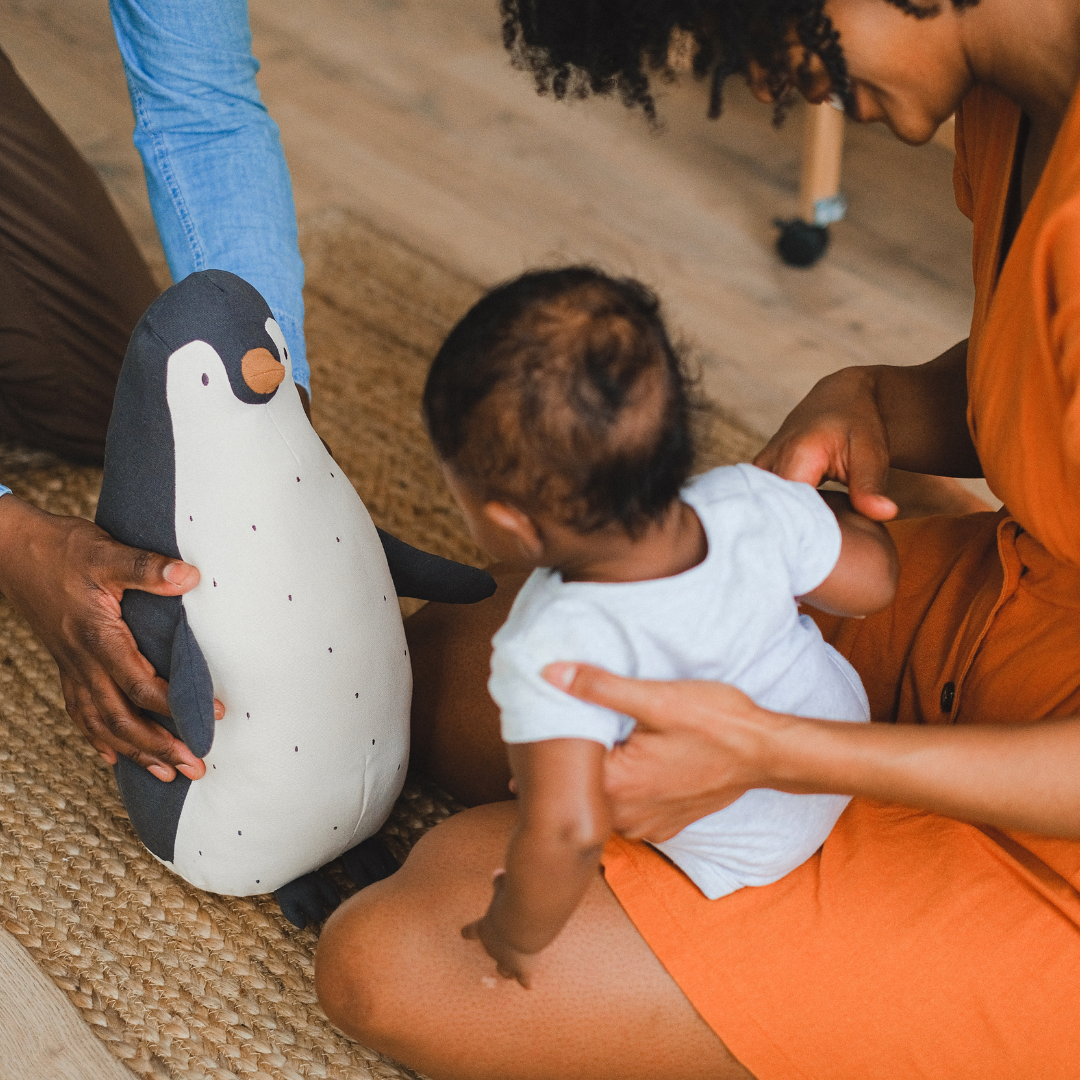 A person holding a toy penguin and a toddler sitting on an adult's lap, with the adult helping the child hold and look at the toy penguin.