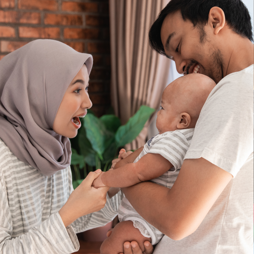 A family with a woman wearing a hijab, a man, and a baby sharing a joyful moment indoors.