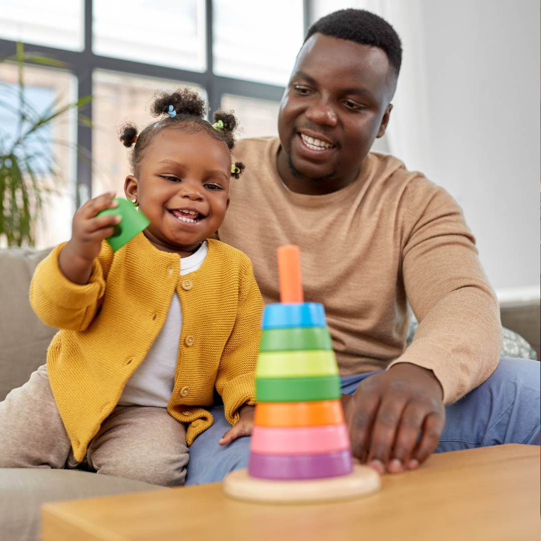 A father and young girl playing with a colorful stacking cone toy indoors, smiling and enjoying quality time.