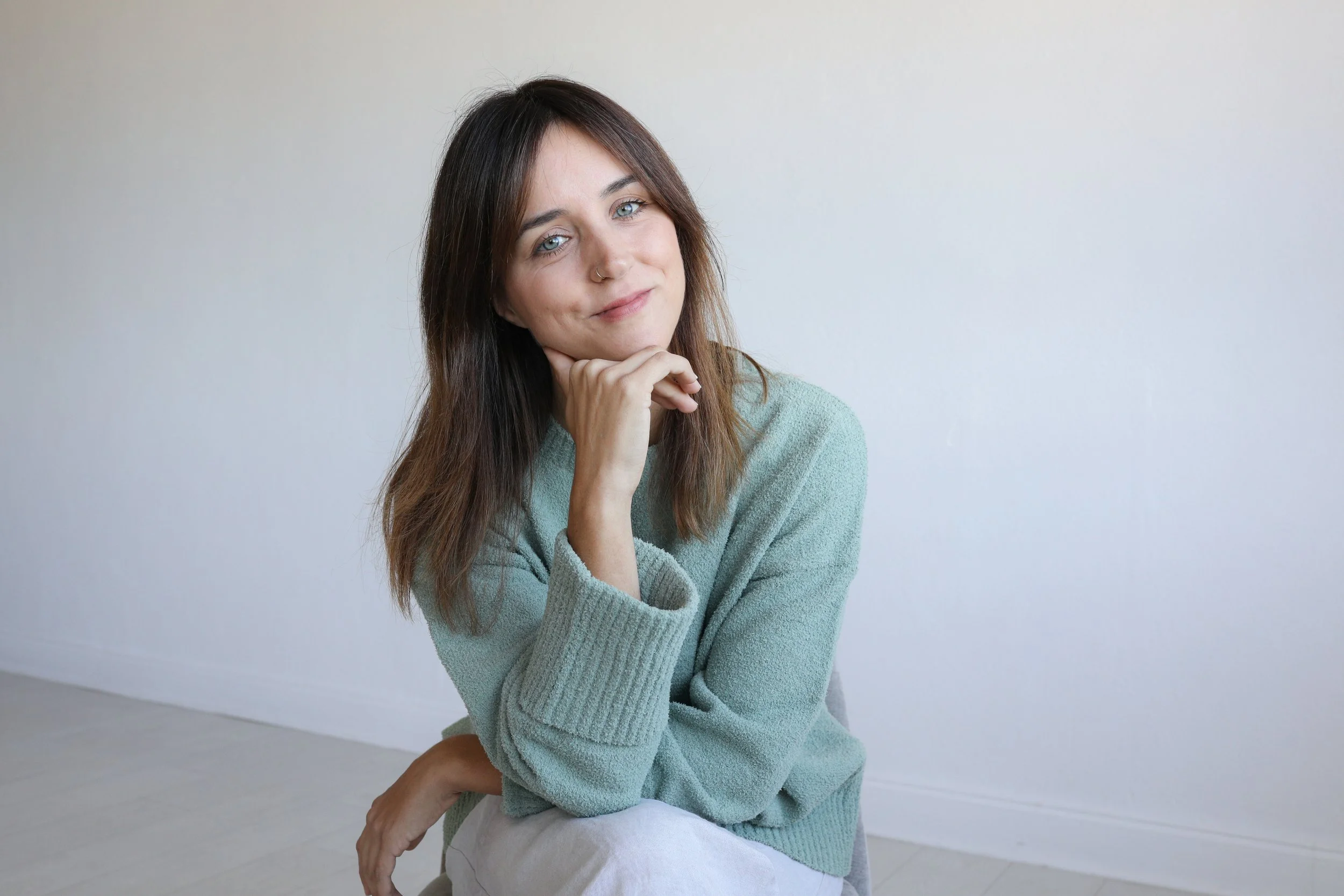 A woman with shoulder-length brown hair, blue eyes, and a nose piercing, wearing a light green sweater, sitting indoors against a plain light-colored wall, resting her chin on her hand and looking at the camera with a slight smile.