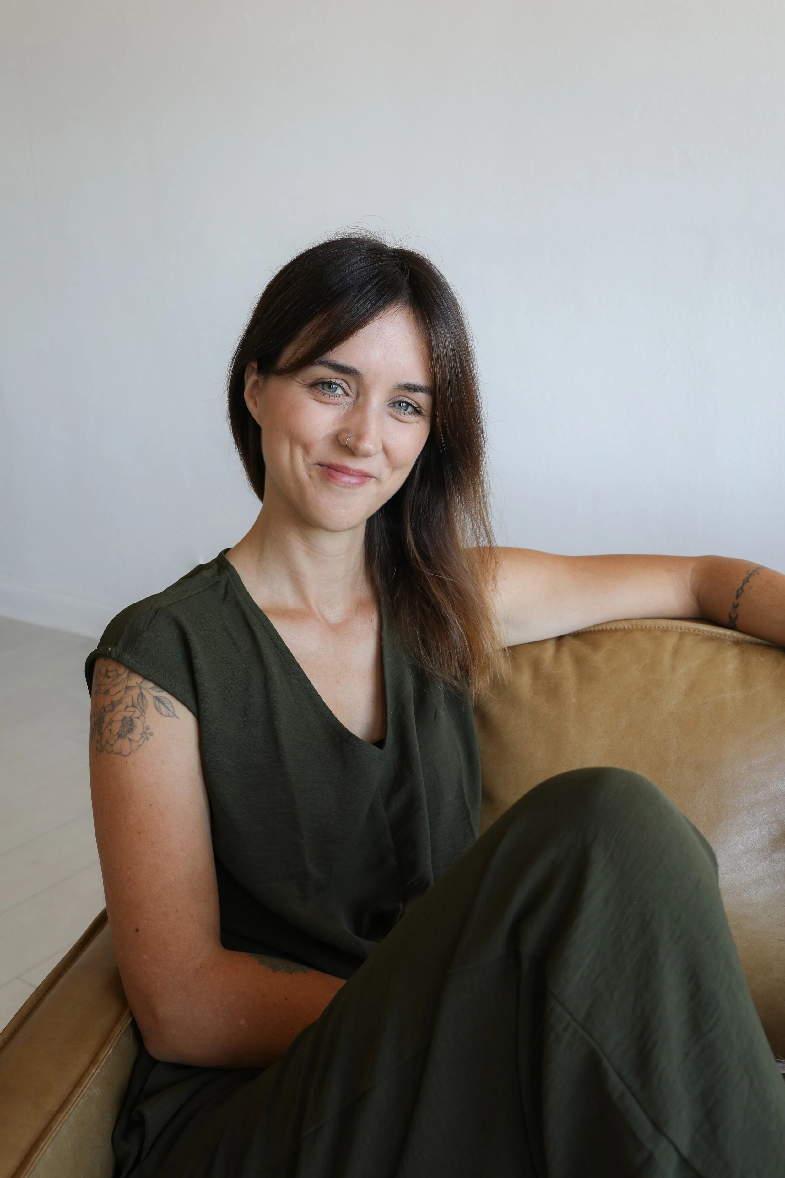 A woman with dark brown hair and tattoos on her arms, wearing a dark green sleeveless top and matching pants, sitting on a tan leather couch and smiling at the camera against a plain white wall.