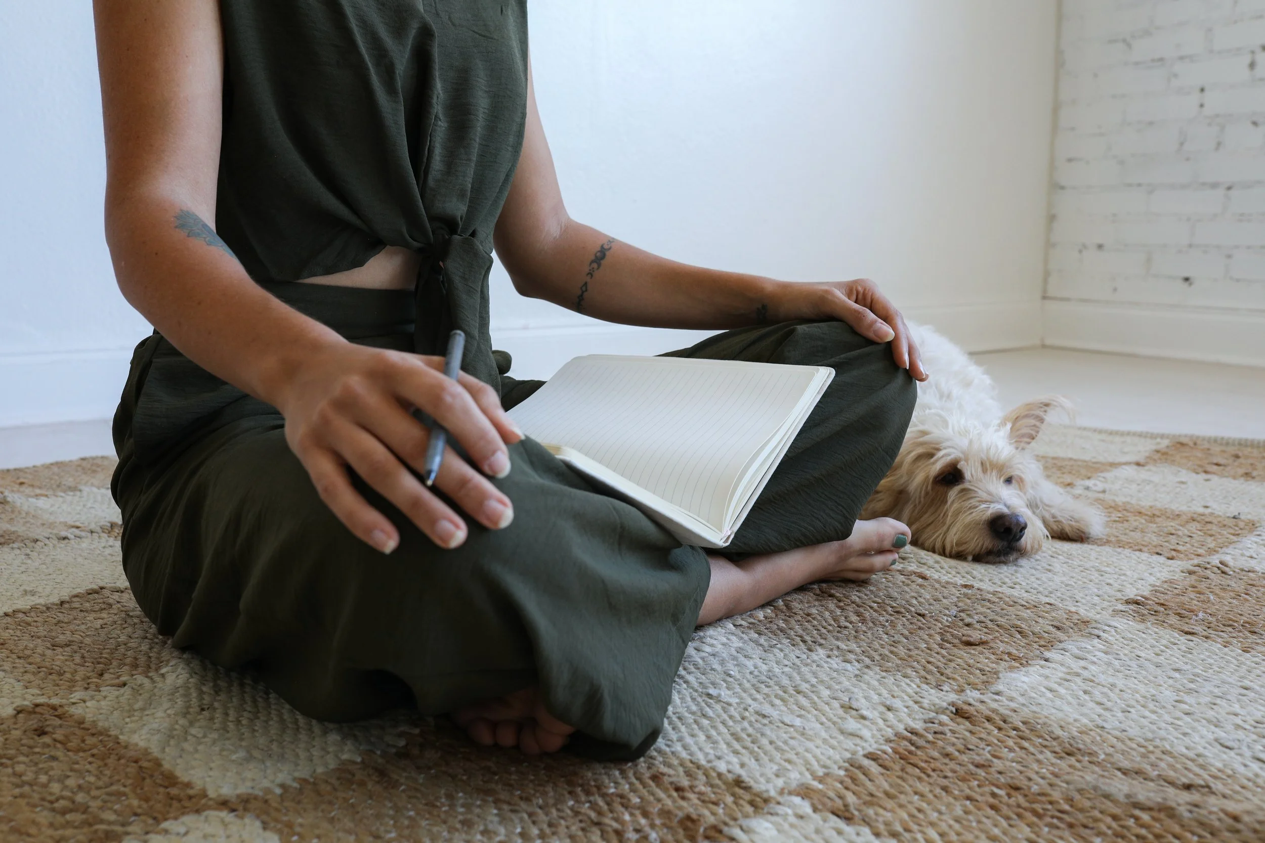 A person sitting cross-legged on a beige patterned rug, holding an open notebook and a pen, with a light-colored dog lying beside them, resting its head on the rug, in a room with white brick walls.