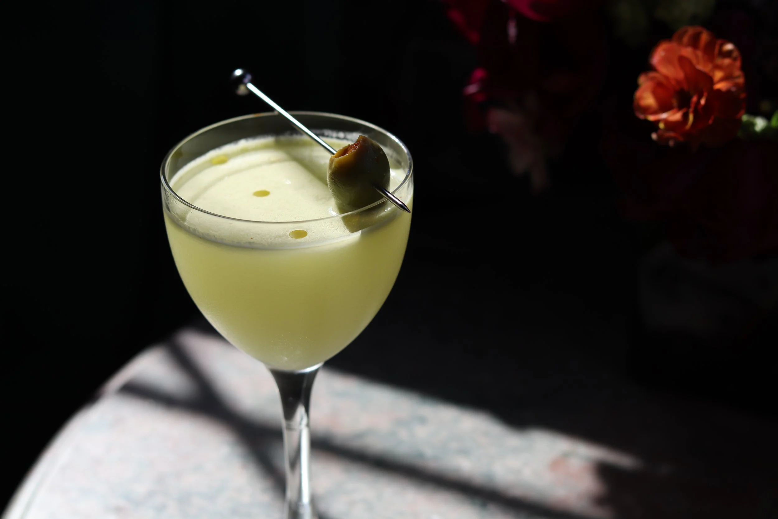 A martini glass filled with a pale yellow cocktail, garnished with a green olive skewered on a cocktail pick.