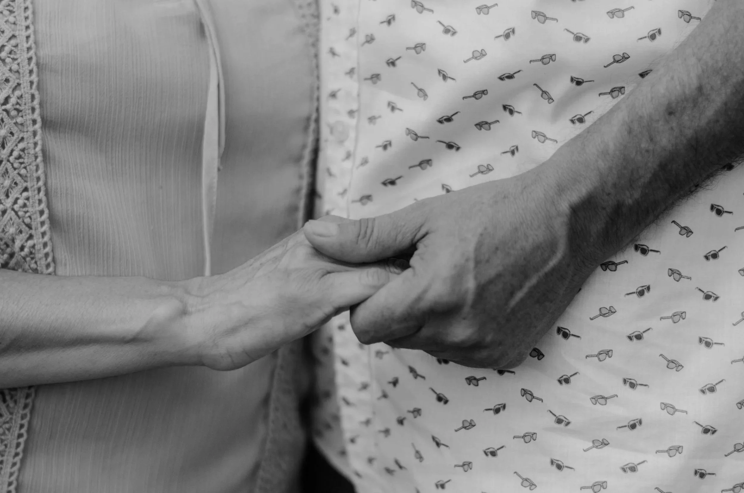 A close-up black-and-white photograph of two people holding hands, one with a lighter skin tone and the other with a darker skin tone, on a bed or fabric surface with a patterned sheet.