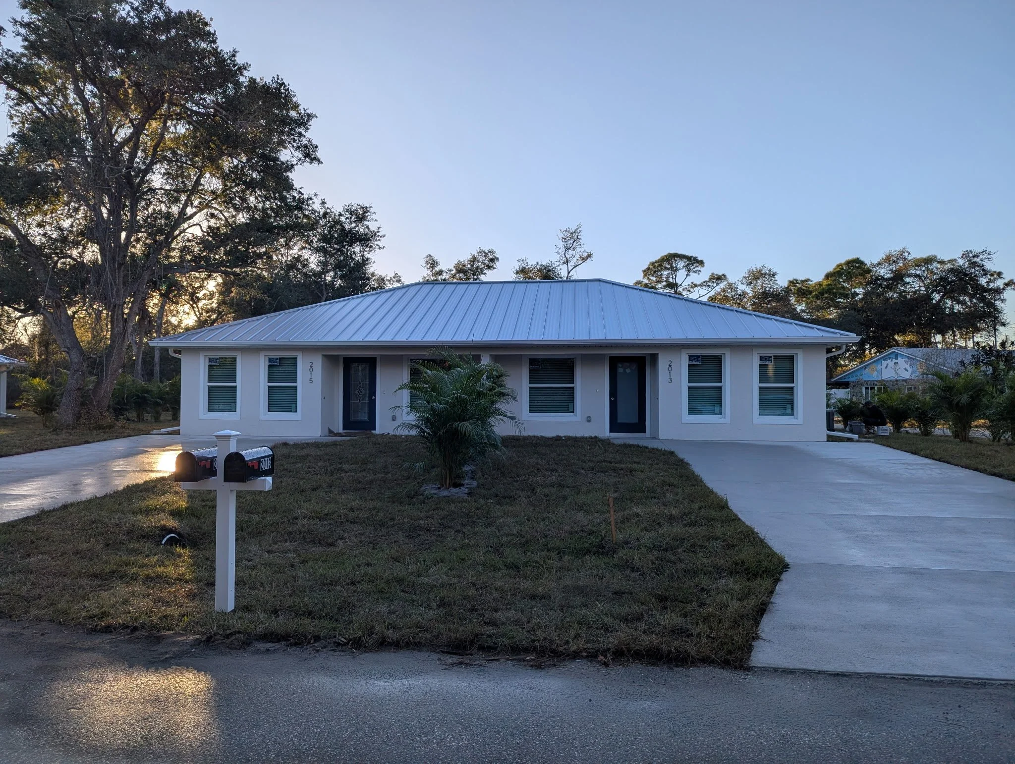 A white single-story house with a metal roof, two front doors, and multiple windows. A driveway runs along the front of the house, and there is a mailbox in the yard. Trees and shrubs surround the house, and the sky is clear.