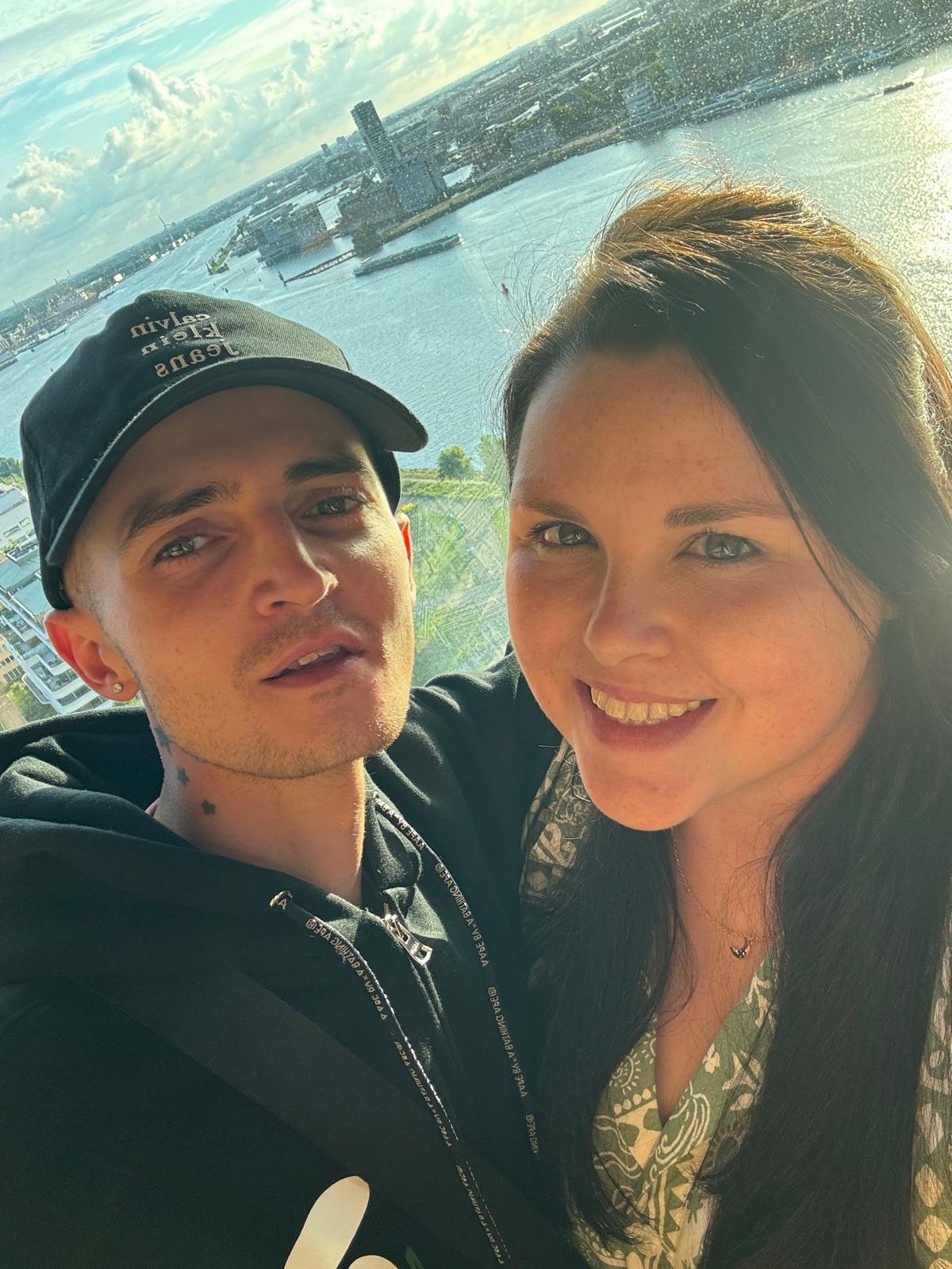 A photo of a smiling woman and a man taking a selfie with a city and river in the background.