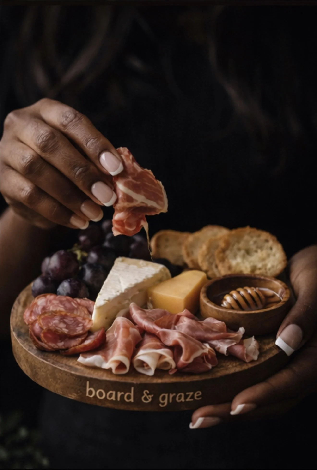 A person holding a cheese and charcuterie board with grapes, cured meats, cheeses, bread, and honey.