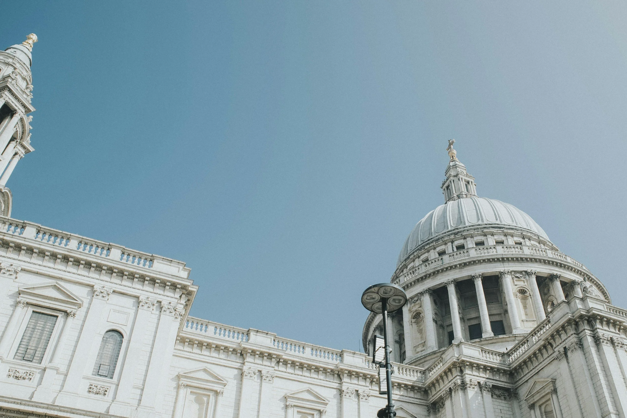 View of St. Paul's Cathedral in London with its large dome, white stone exterior, and classical architectural details against a blue sky. A street lamp is also visible in the foreground.