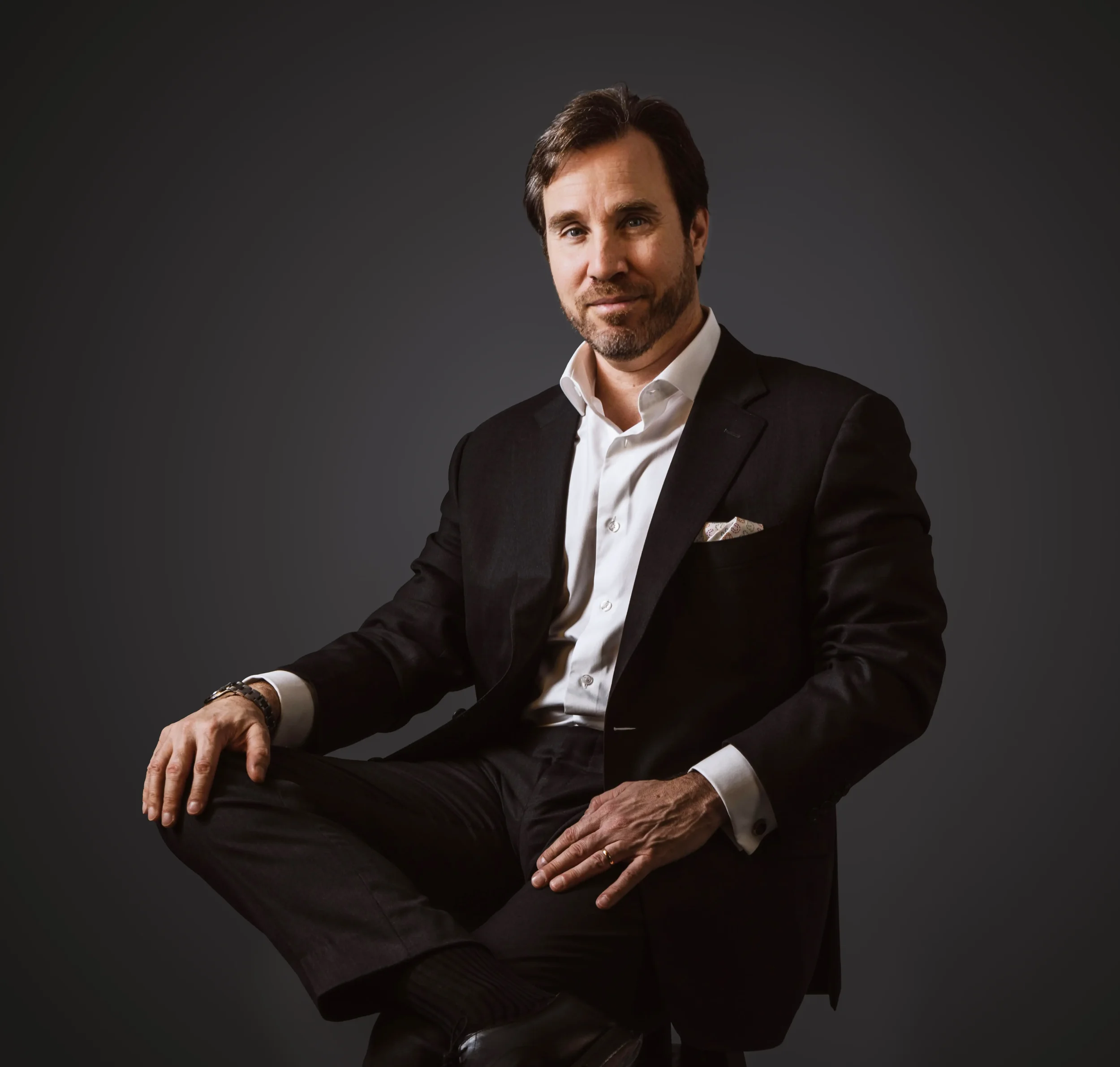 Homem de terno preto, camisa branca, cabelo castanho, barba e expressão confiante sentado contra fundo escuro.