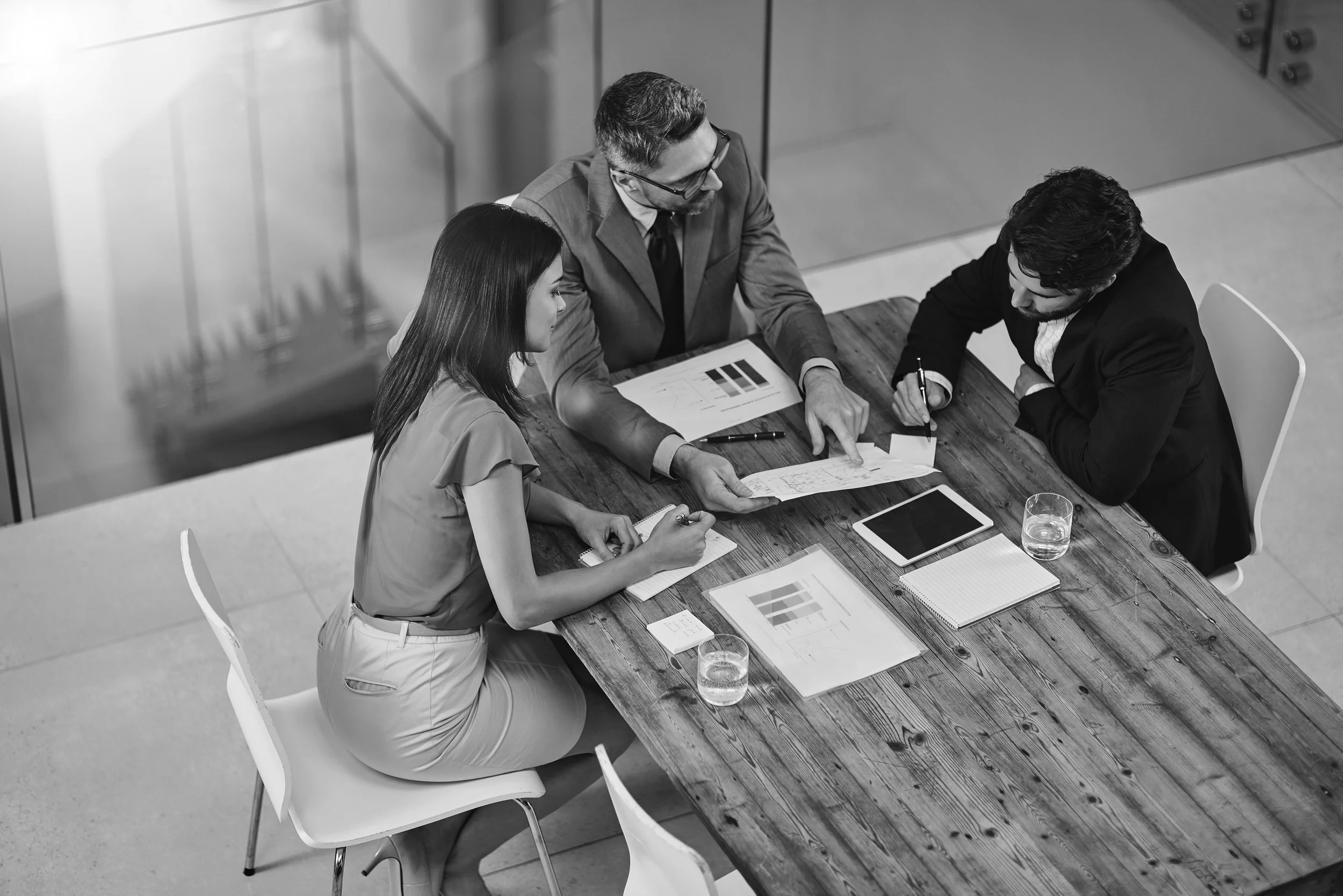 Três pessoas em uma reunião de negócios ao redor de uma mesa de madeira, analisando gráficos e documentos, com uma xícara de água e um tablet na mesa.
