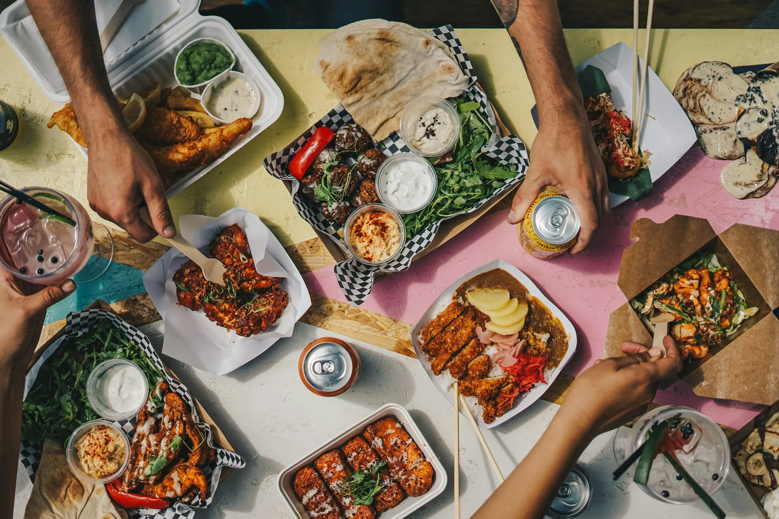 Top-down view of a colorful table filled with various dishes including fried chicken, fries, sauces, salads, rolls, and beverages, with multiple hands reaching for food and drinks.