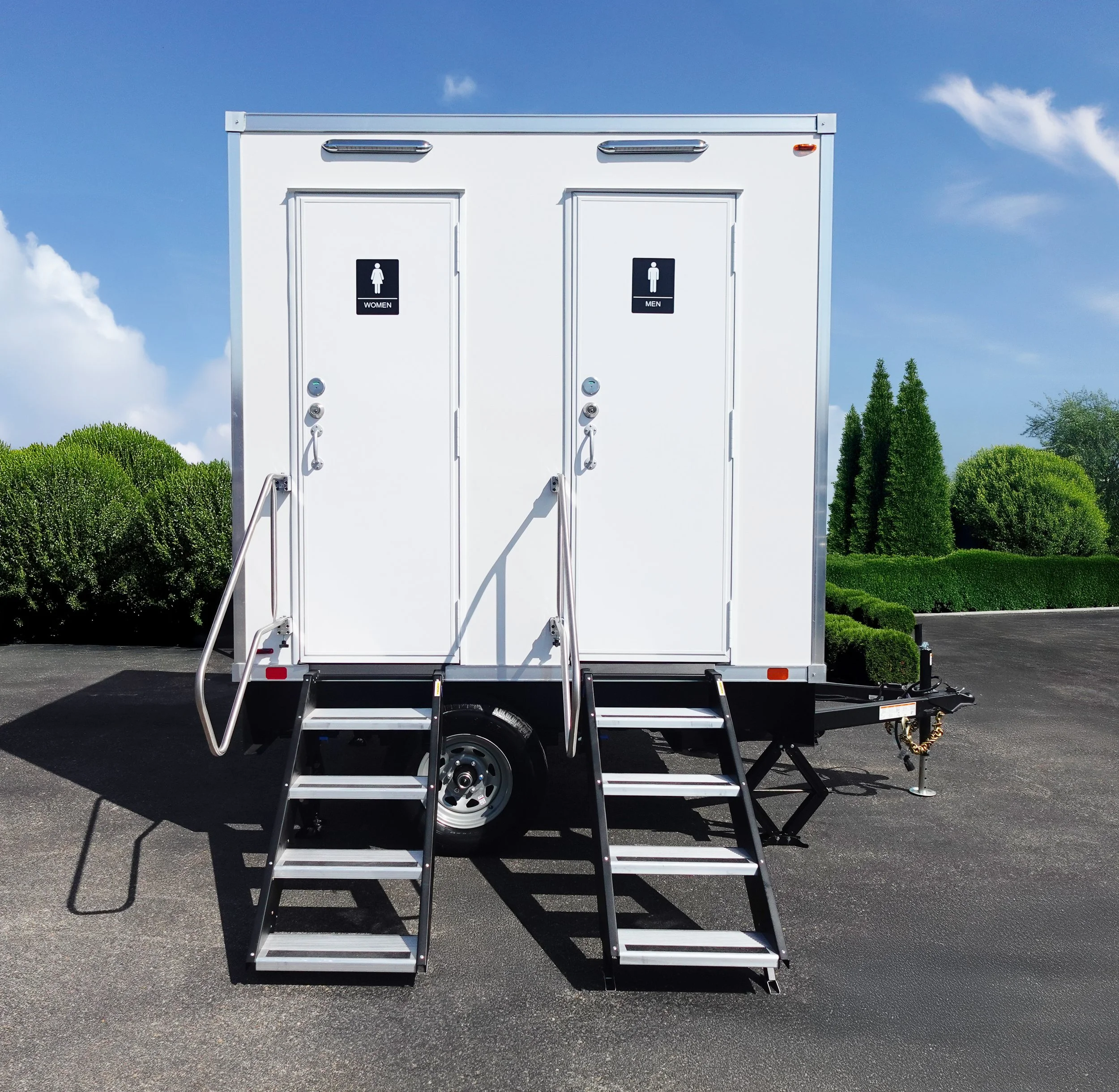 Portable restroom trailer with separate entrances for women and men, positioned on asphalt with green bushes and trees in the background under a blue sky.