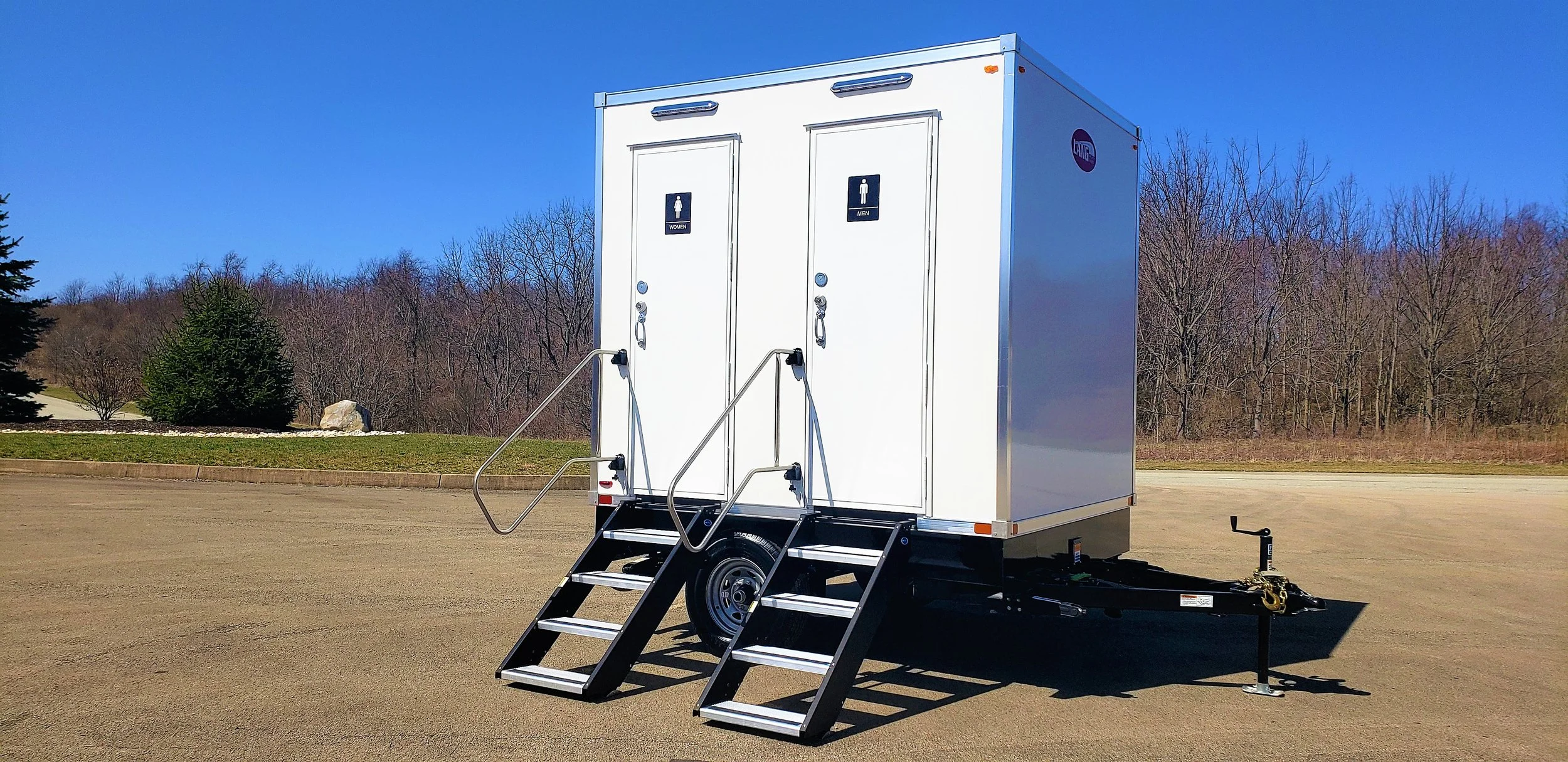 A portable restroom trailer with separate doors for women and men, set on an open paved area with trees and clear blue sky in the background.