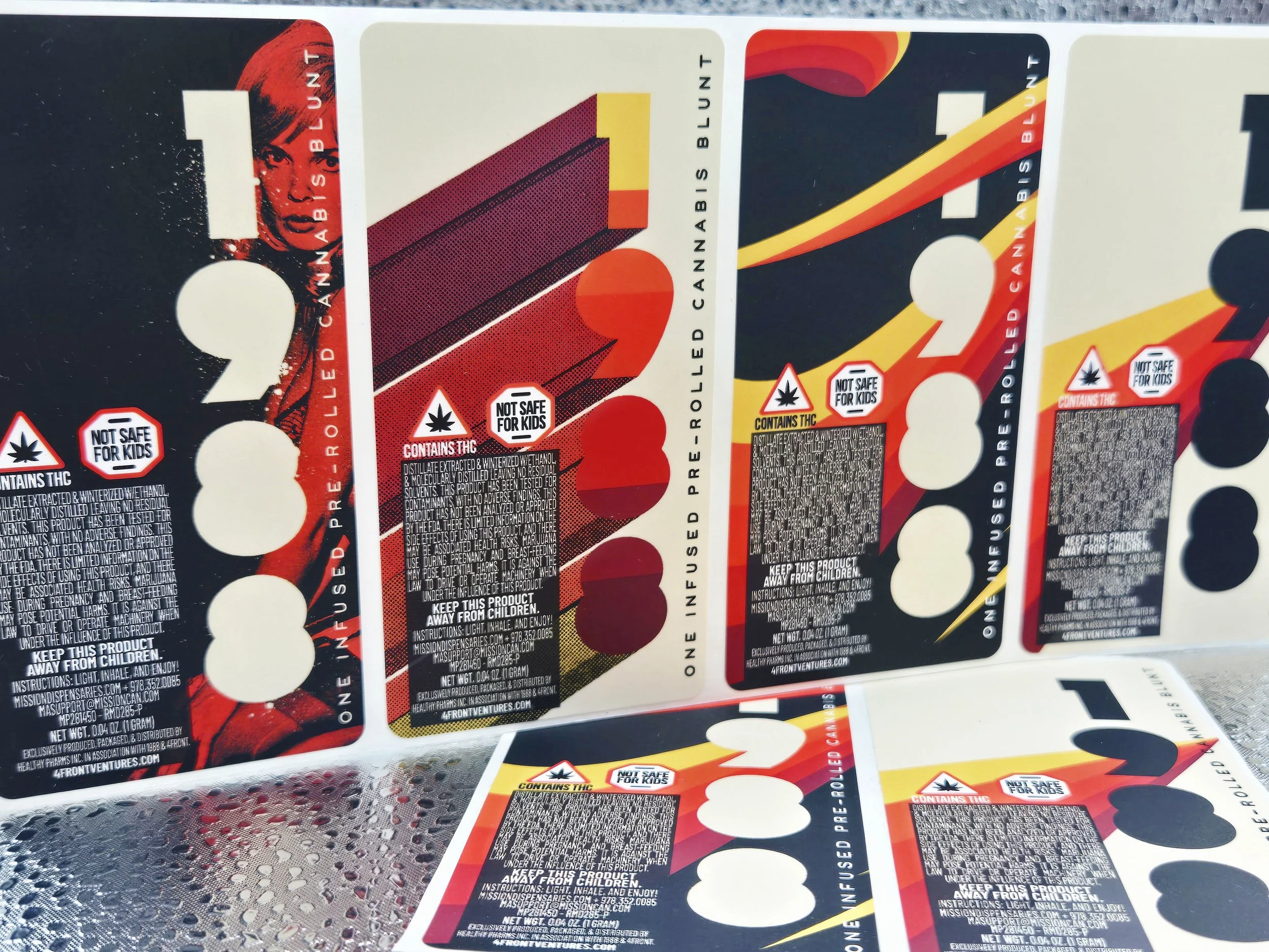 Multiple cannabis product labels with red, yellow, and black designs, featuring cautions about THC content and safety warnings, displayed on a metallic surface.