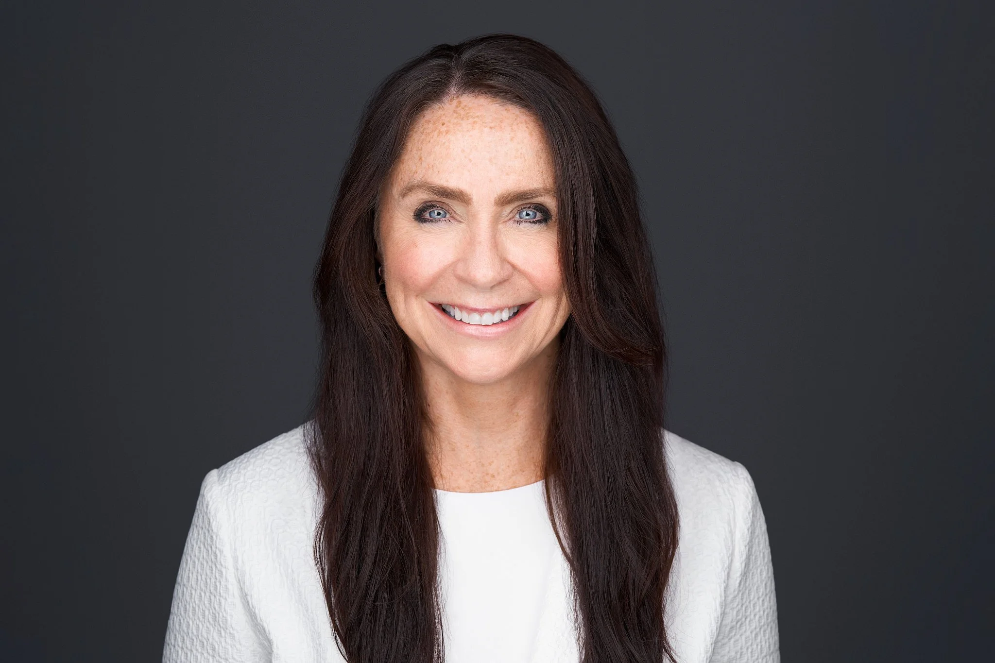 A smiling woman with long dark hair and blue eyes against a dark gray background.
