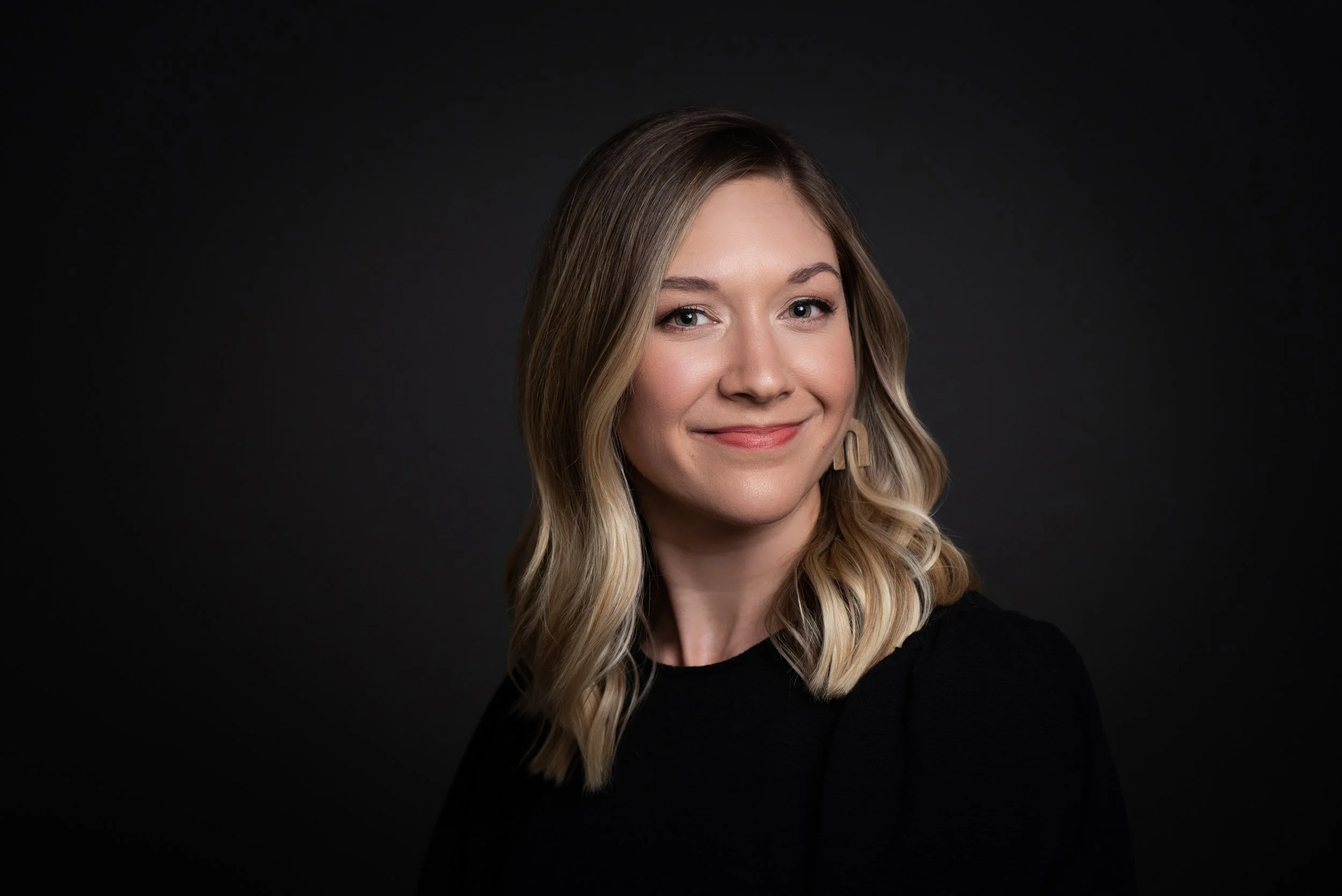 A woman with shoulder-length wavy blonde hair, wearing a black top, smiling and looking at the camera against a dark background.