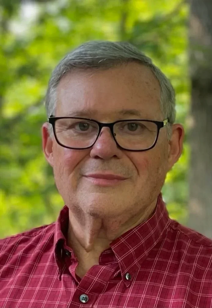 Portrait of an older man with glasses, short gray hair, wearing a red plaid shirt, outdoors with green foliage in the background.