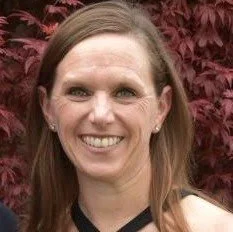 A woman with long, light brown hair smiling in front of a background of red leaves.