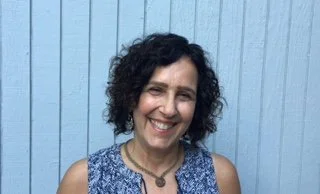 A woman with curly black hair smiling in front of a light blue wooden wall.