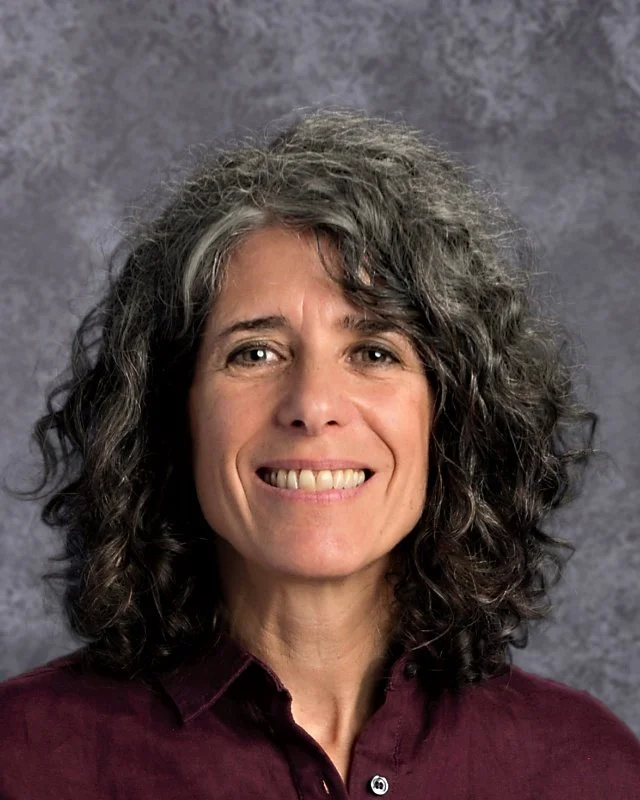 A woman with shoulder-length curly salt-and-pepper hair, smiling, wearing a dark red collared shirt, with a neutral gray textured background.