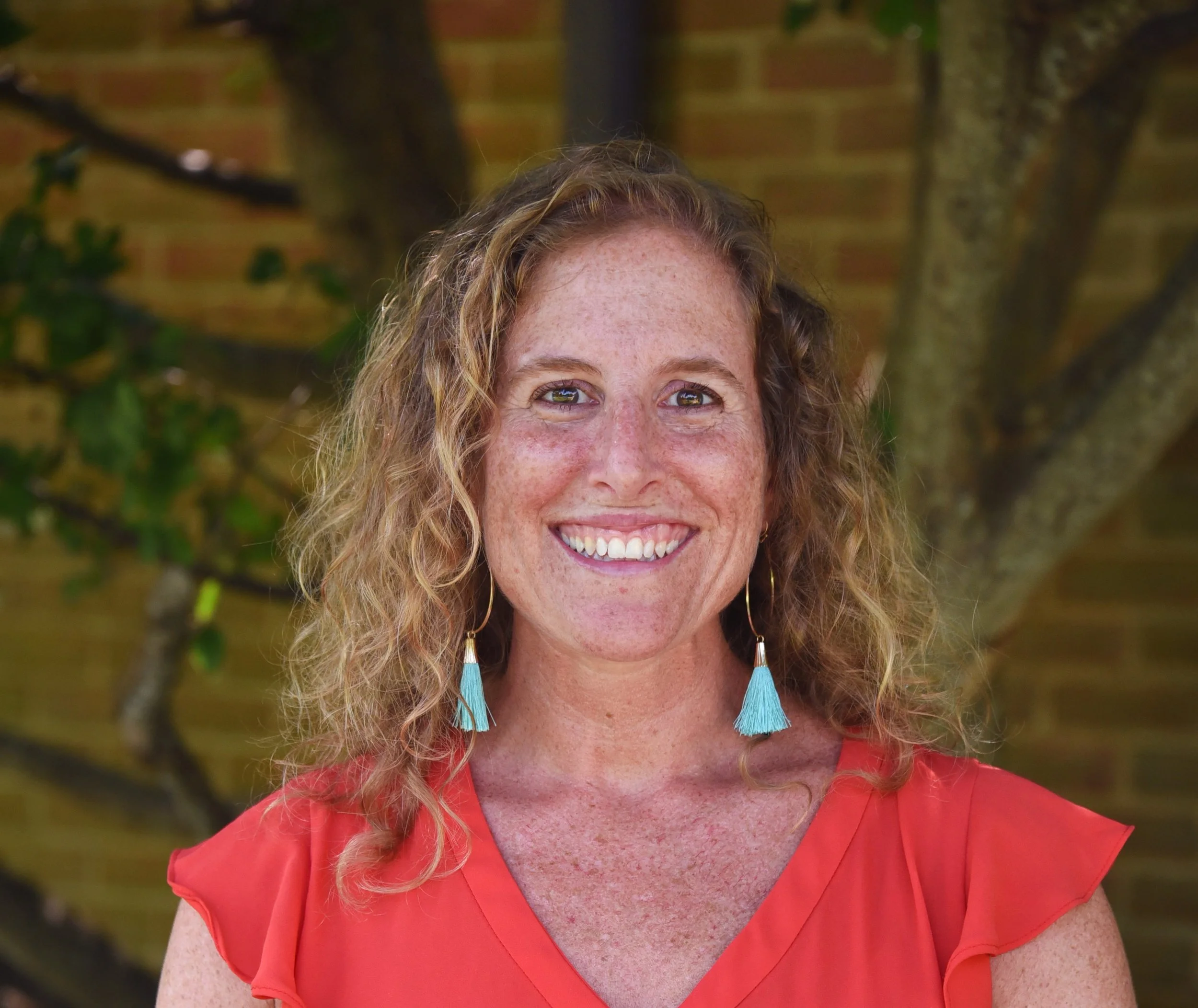 A woman with curly red hair, freckles, and a bright smile, wearing earrings with blue tassels and an orange top, standing outdoors with a tree and brick wall in the background.