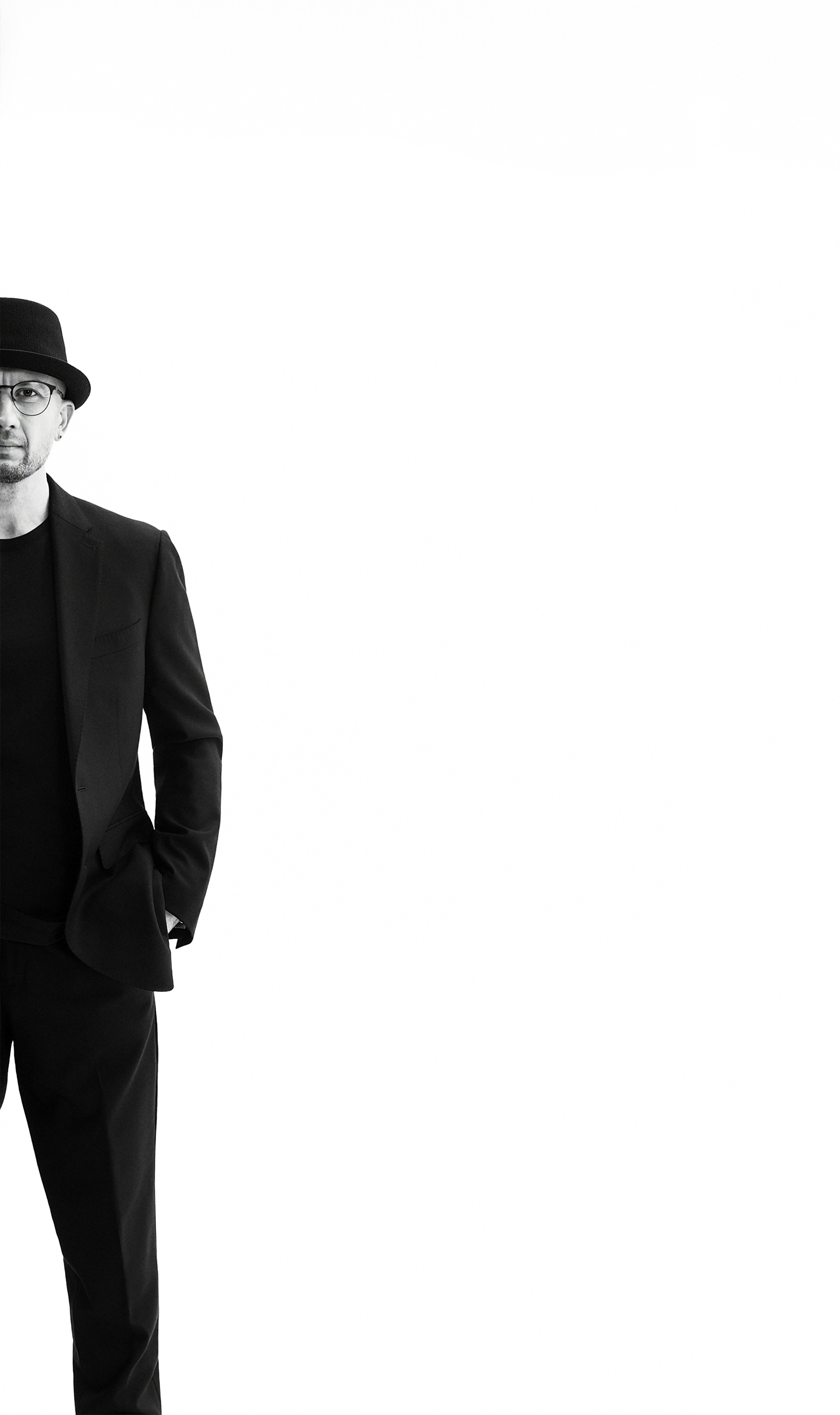 A man dressed in a black suit, black T-shirt, and wearing glasses and a black hat, standing with his left hand in his pocket against a white background.