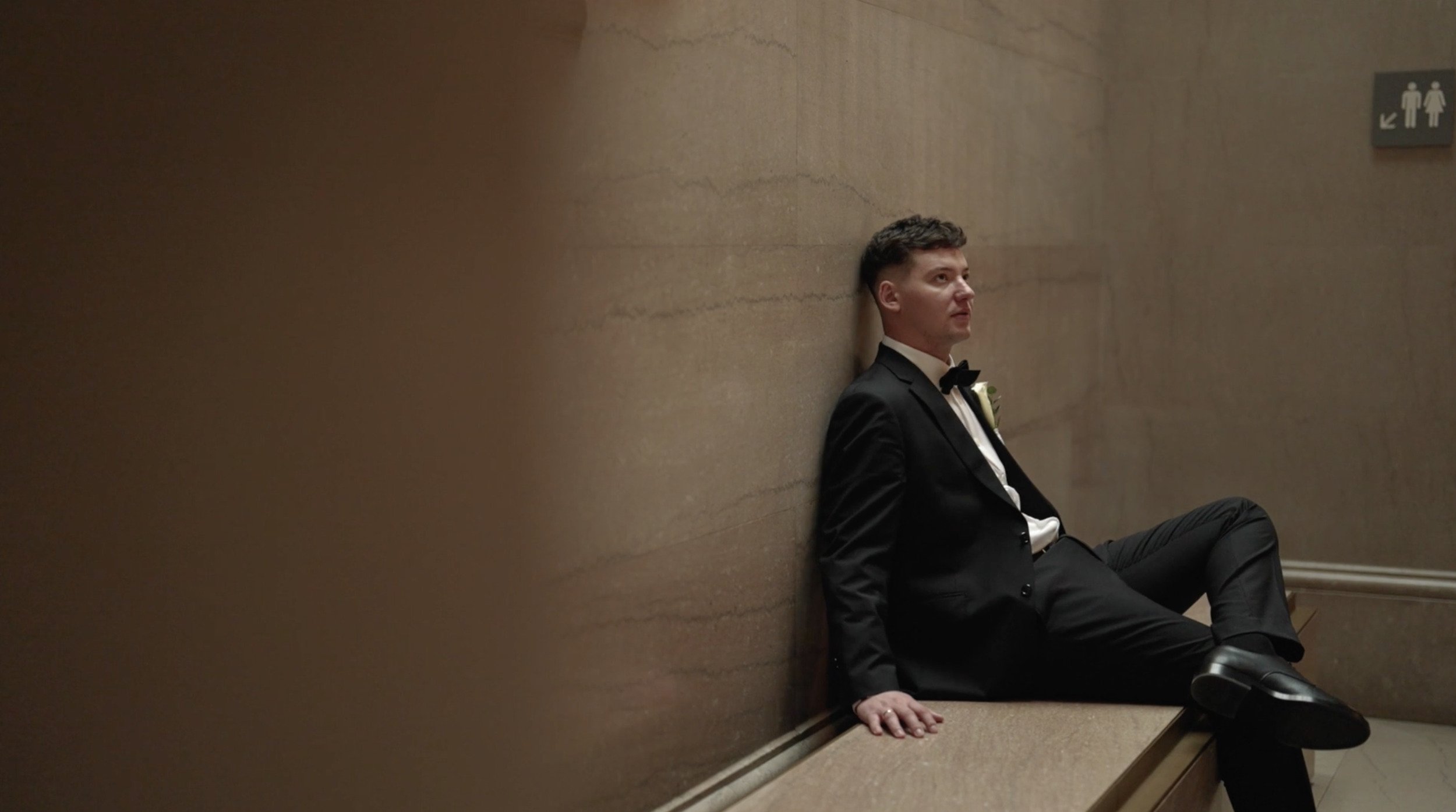 A man in a tuxedo sitting against a wall in a stairwell with a contemplative expression.