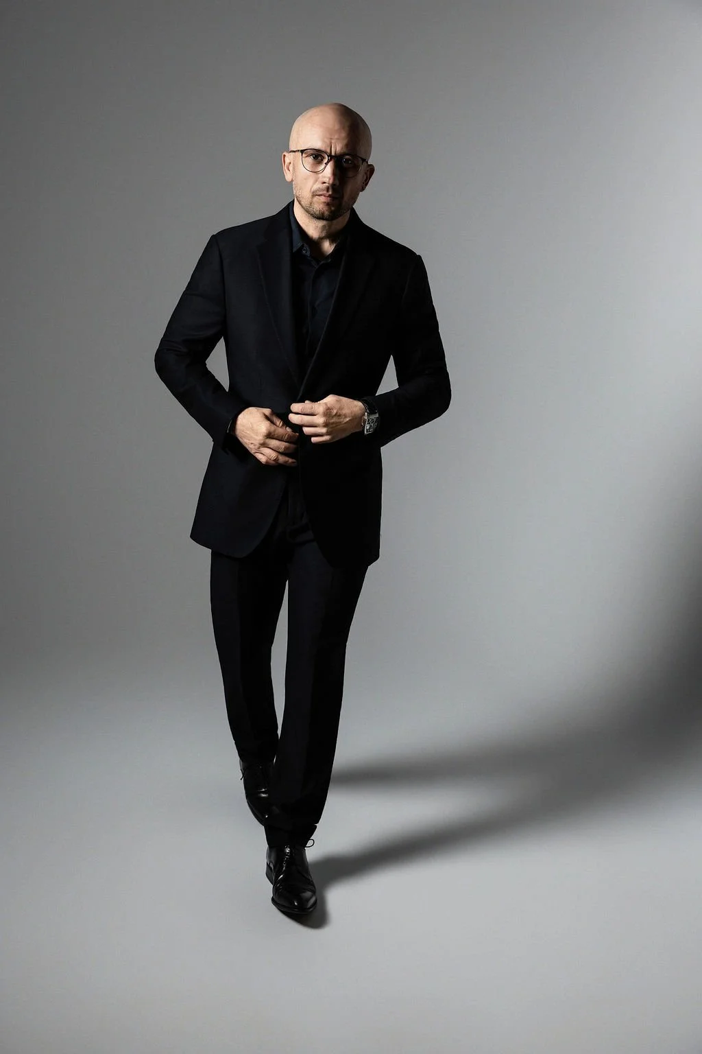 A bald man wearing glasses in a black suit adjusts his jacket while standing against a plain gray background.