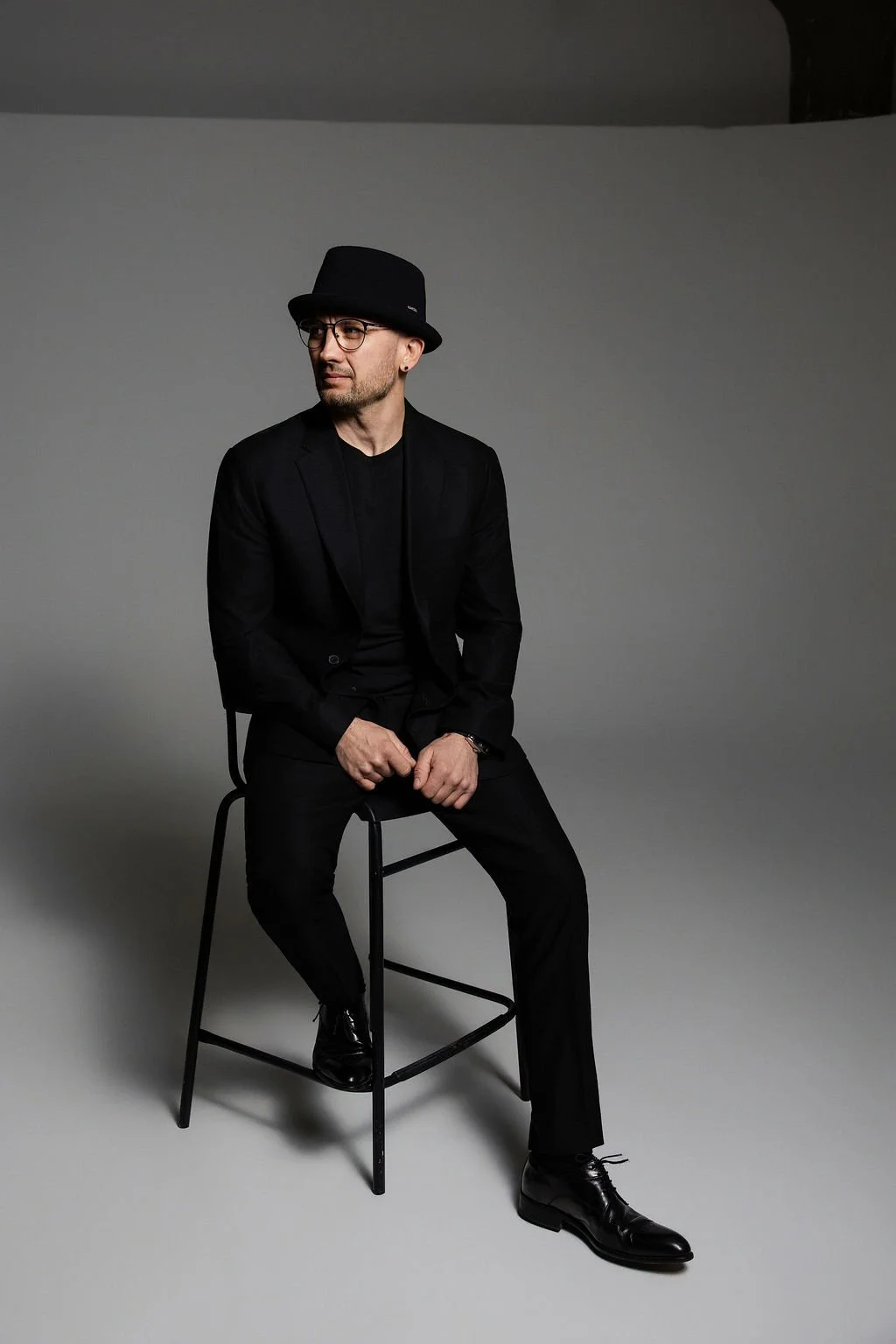A man dressed in black with a black hat, glasses, and beard, sitting on a stool against a plain grey background.