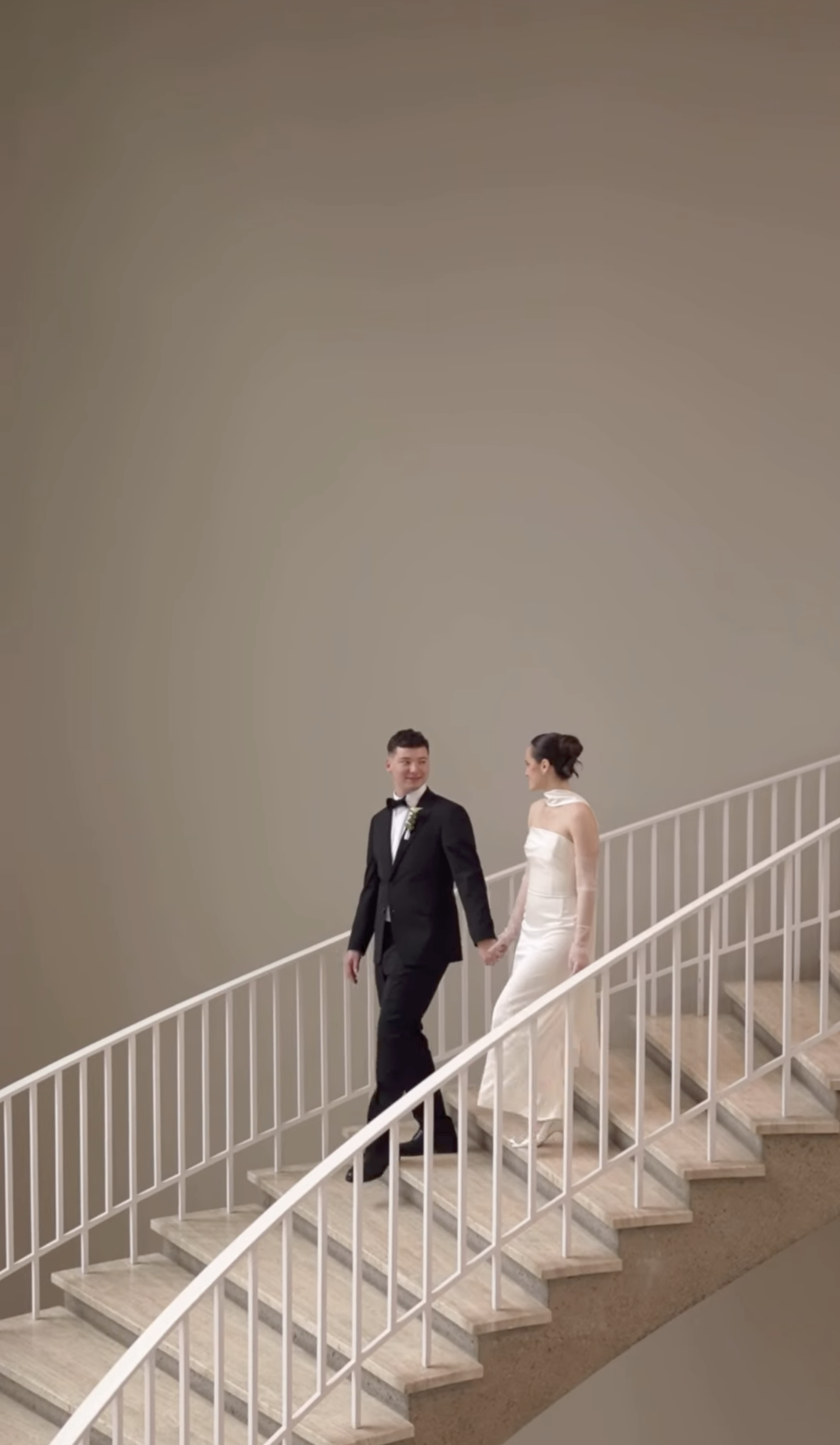 A newlywed couple holding hands and walking down a staircase, the groom in a tuxedo and the bride in a white wedding dress.