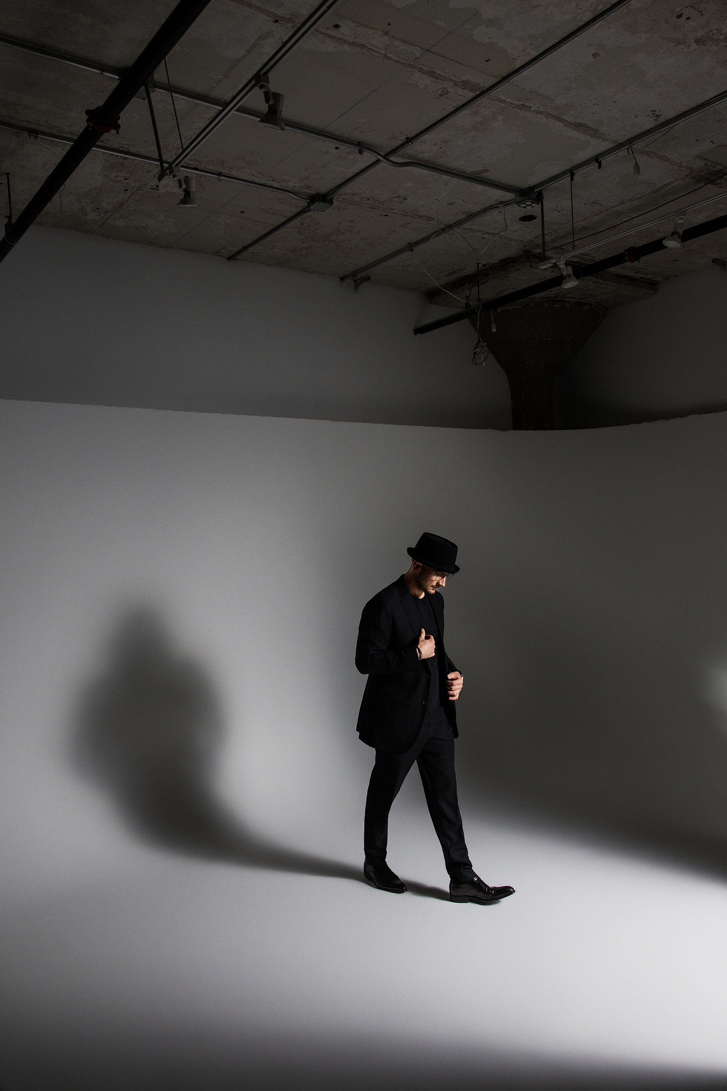 A man in a black suit and hat standing on a grey studio backdrop, looking down with hands adjusting his jacket, casting a shadow on the background.