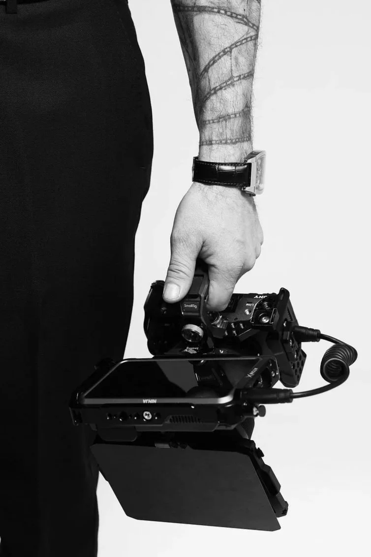 A person holding a camera with a monitor attached, wearing a watch and showing a tattooed arm, in black and white photograph.