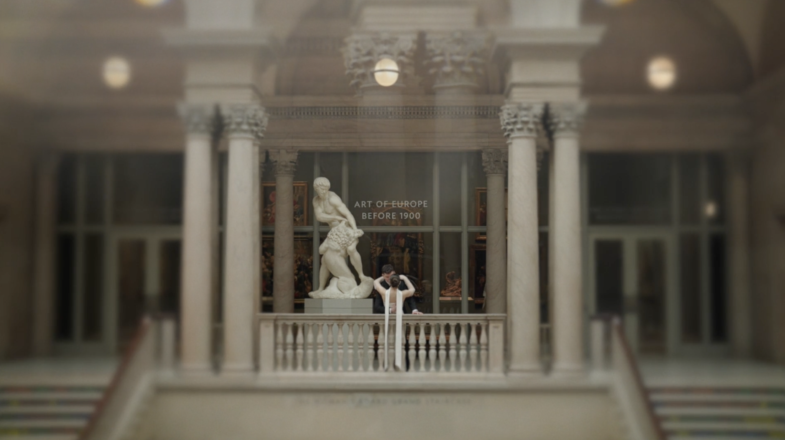 Inside a museum with classical architecture, displayed sculptures, and framed paintings. Two visitors are taking photos in front of a large statue. A sign reads 'Art of Europe Before 1900'.