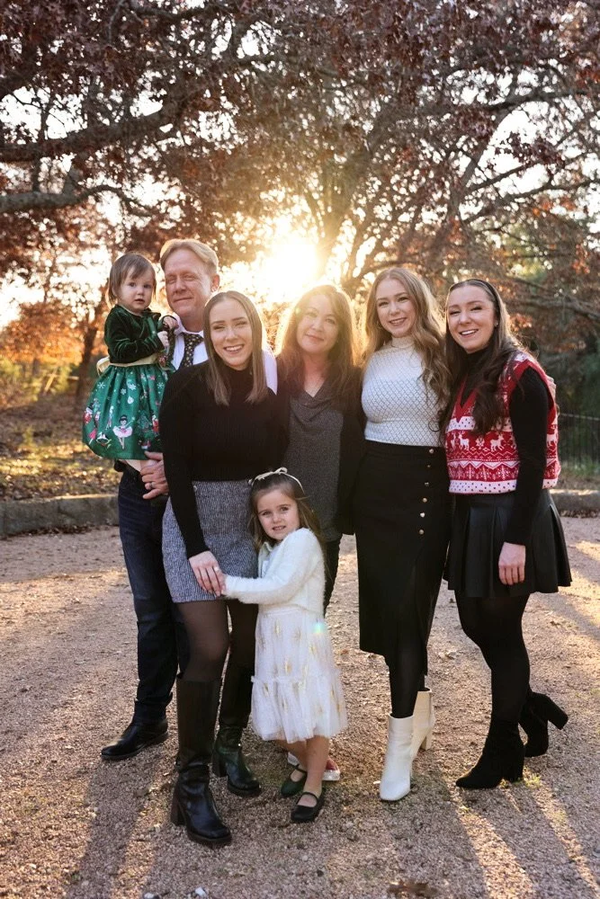 Family group outdoors at sunset, including three adult women, one man, and two young girls, standing on a gravel surface with trees in autumn foliage in the background.