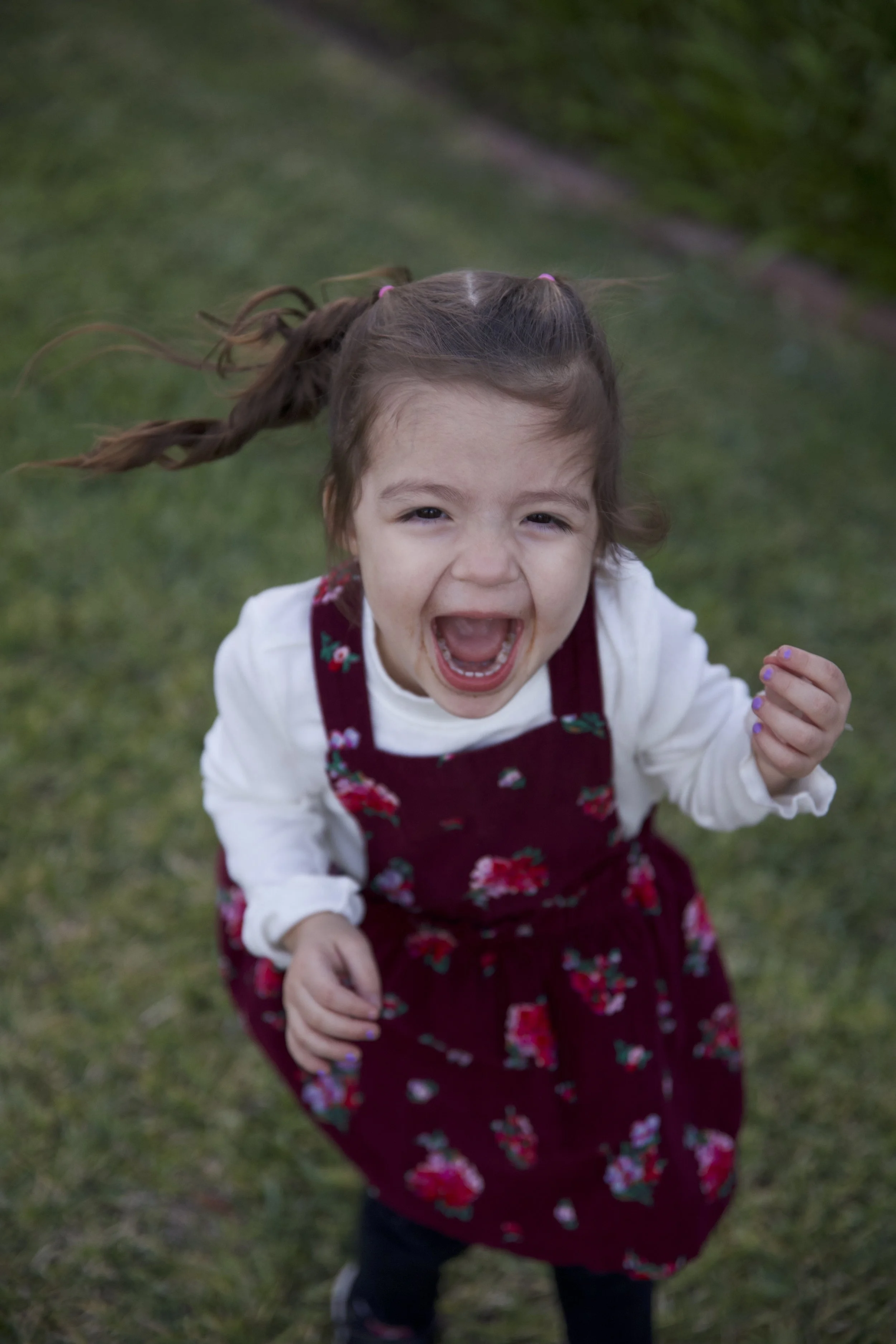 A young girl with braided hair and a flower-patterned dress is smiling and running outdoors on a grassy area.
