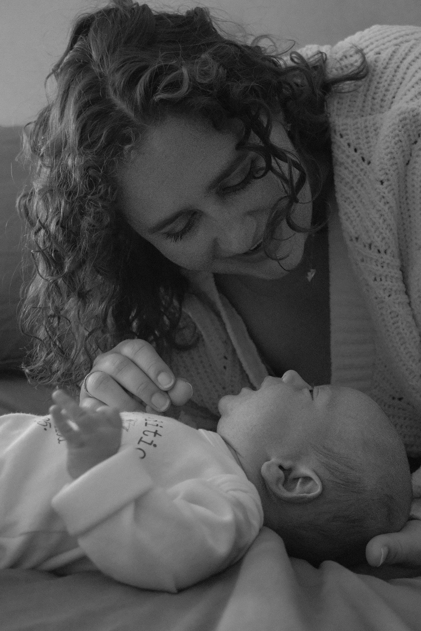 A woman with curly hair gently touching a baby's face while lying together, both looking at each other lovingly.