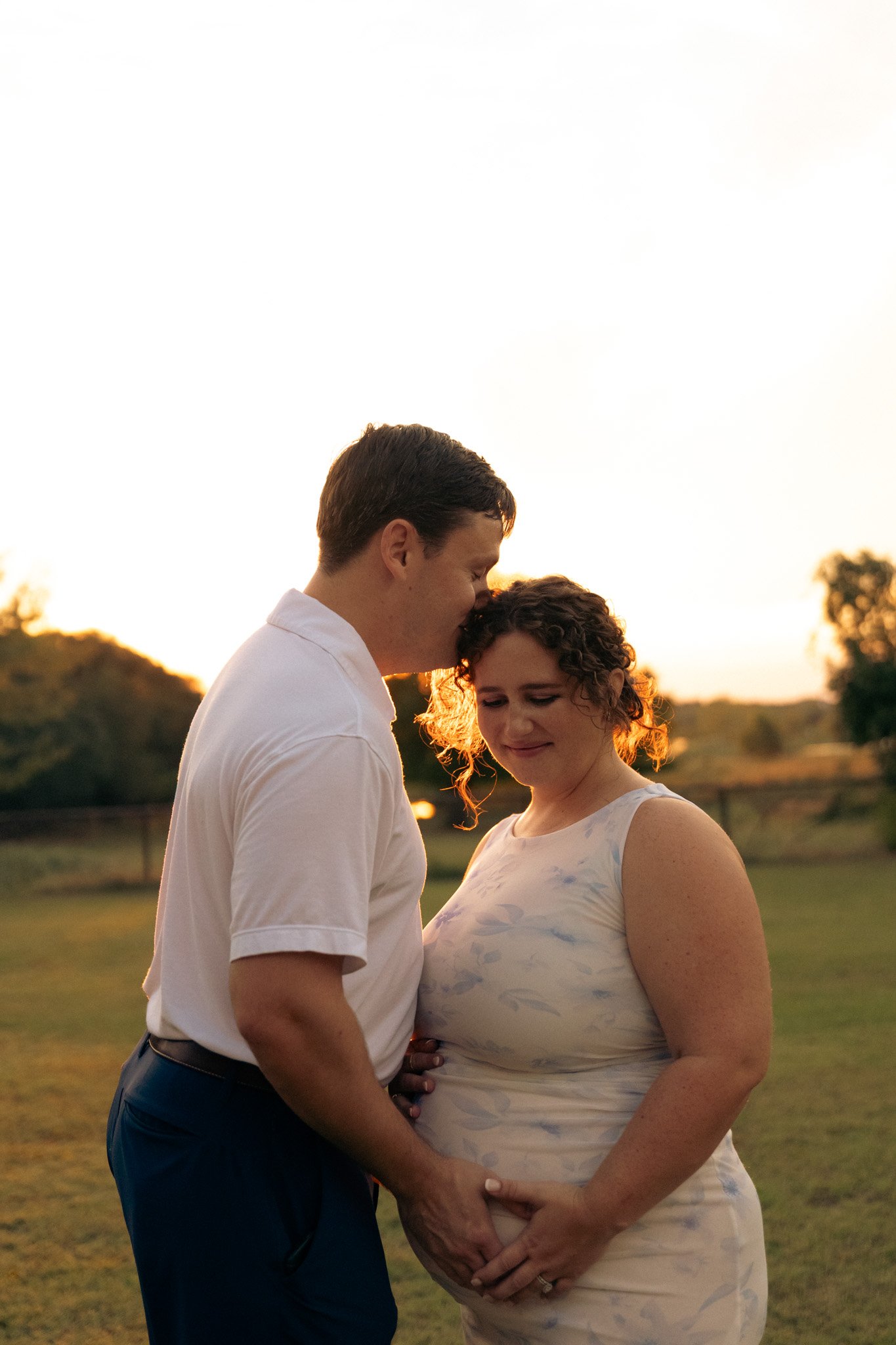 A pregnant woman with curly hair in a white dress is holding her baby bump while a man in a white shirt and dark pants kisses her on the forehead during sunset outdoors.