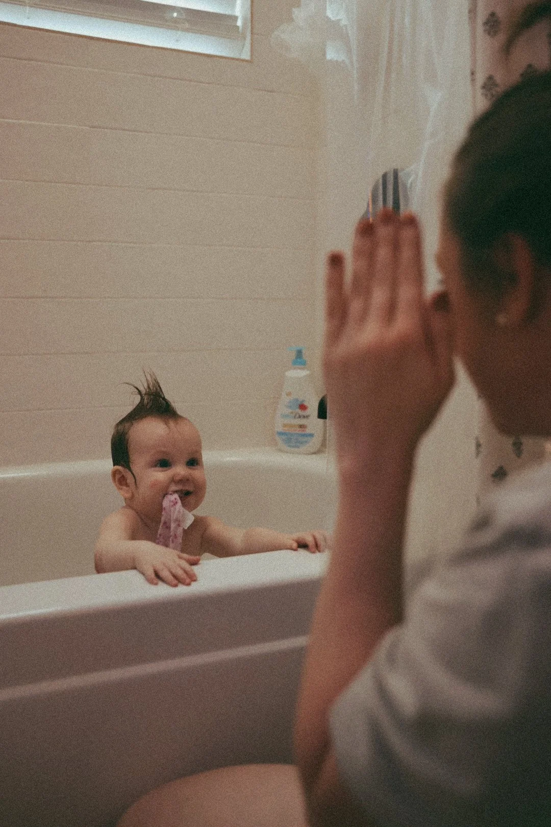 A baby with wet hair and a cloth in their mouth smiling in a bathtub, looking at an adult with hands near their face, in a bathroom with beige walls and a bottle of soap or shampoo in the background.