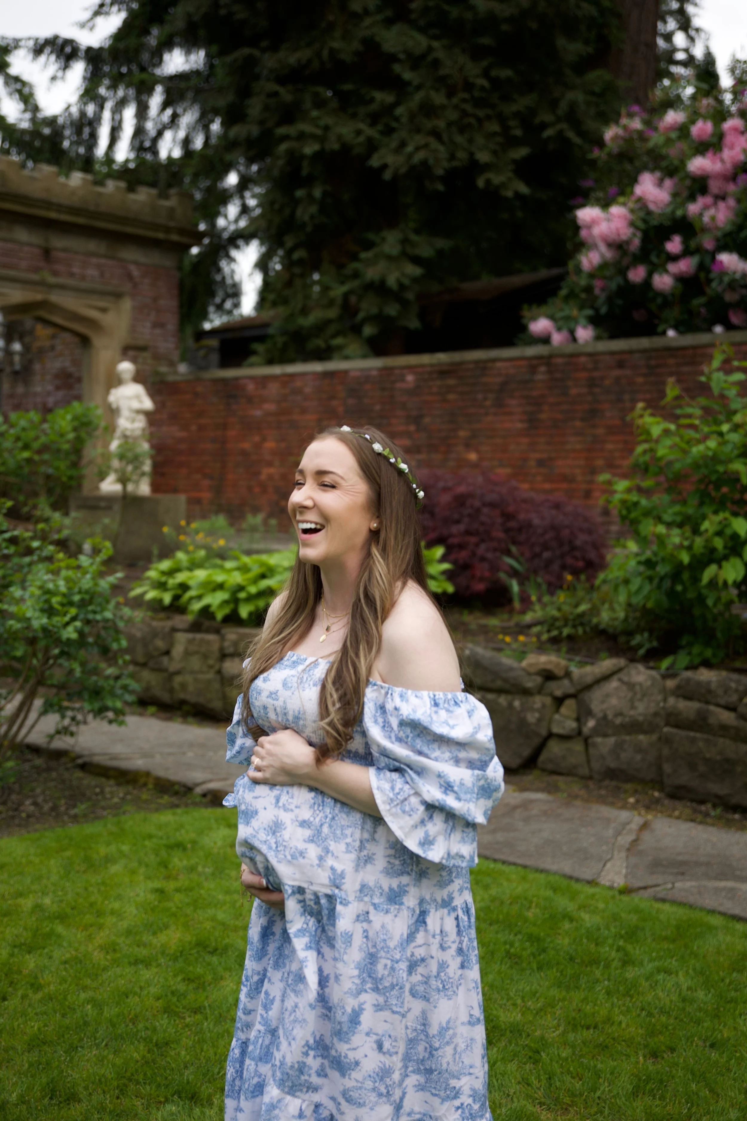 A woman in a blue and white off-shoulder dress with floral patterns, smiling and holding her pregnant belly in a garden setting with lush greenery, colorful bushes, a brick wall, and a statue in the background.