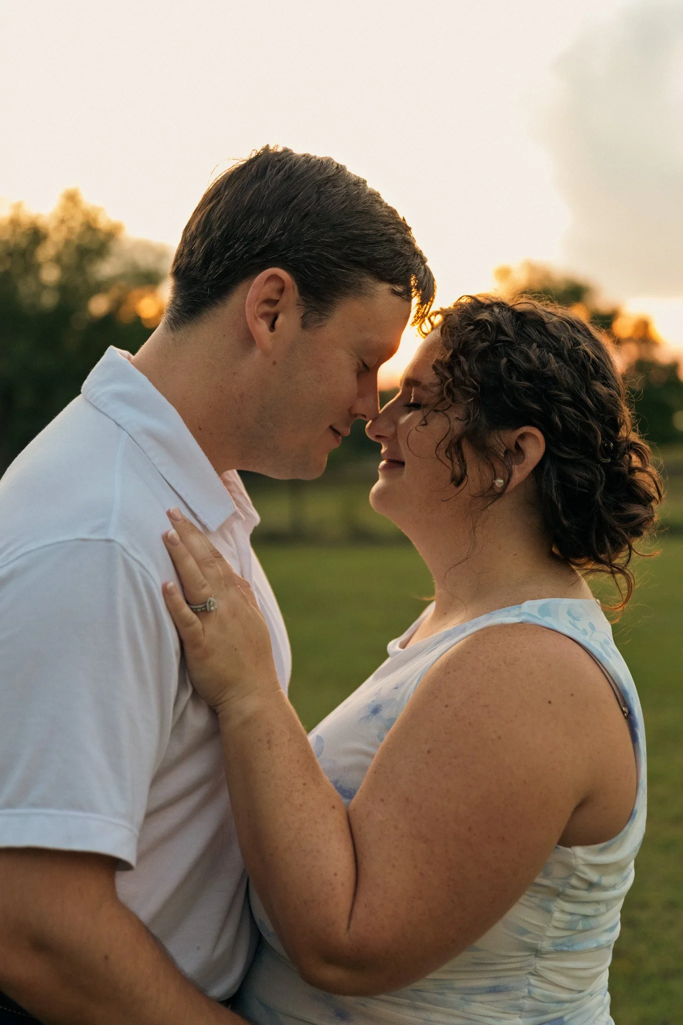 A couple with their foreheads touching, smiling in a park during sunset.