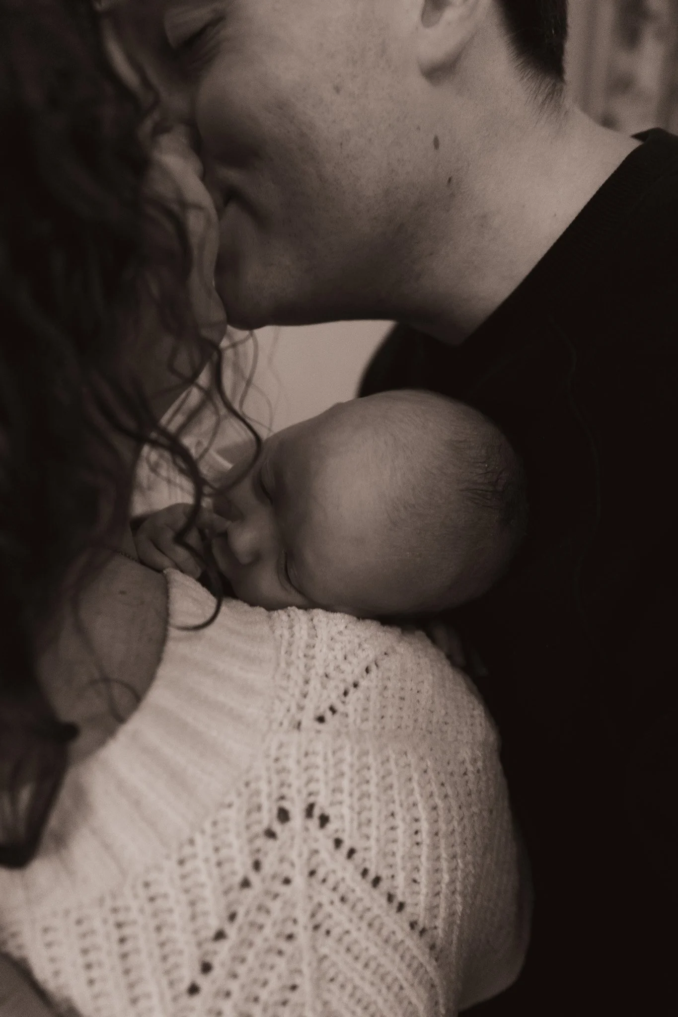 A black and white photo of a man and woman kissing, with a sleeping baby tucked between them.