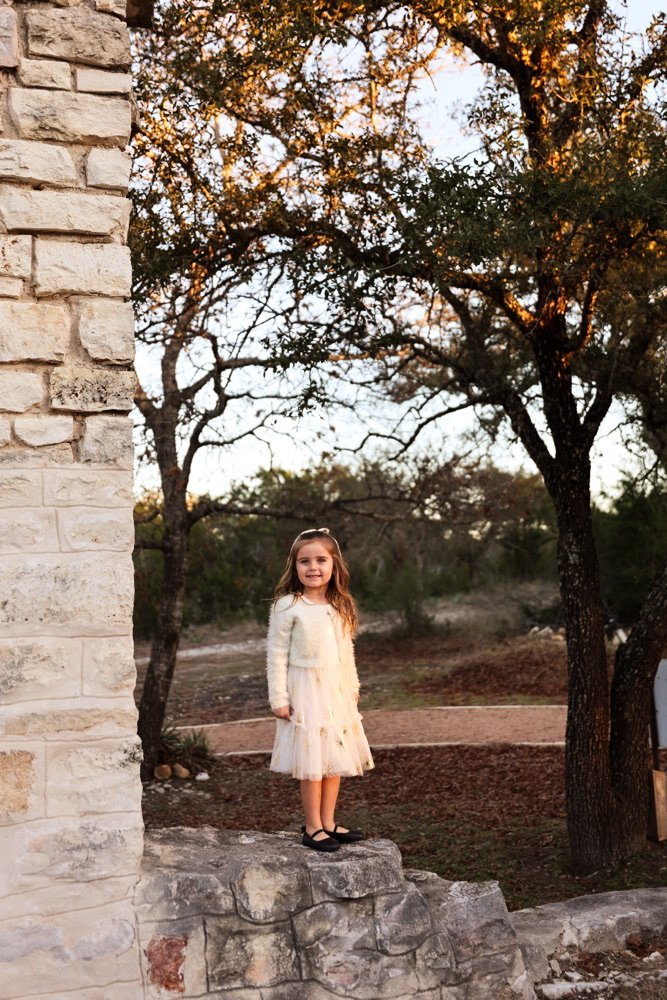 A young girl with long hair, wearing a white dress and black shoes, standing on a stone ledge outdoors near trees at sunset.