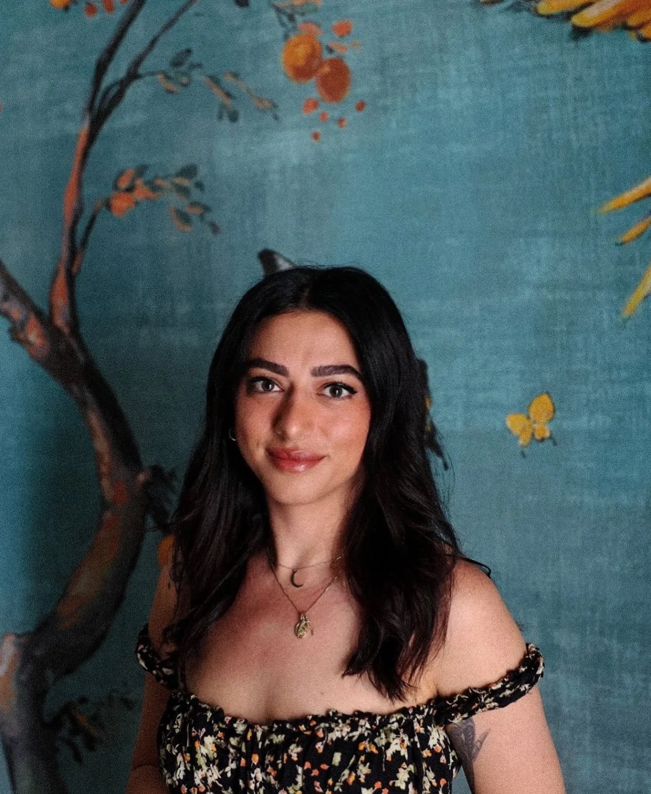 Portrait of a young woman with dark wavy hair, wearing an off-shoulder floral dress and layered necklaces, standing in front of a colorful mural depicting trees and butterflies.