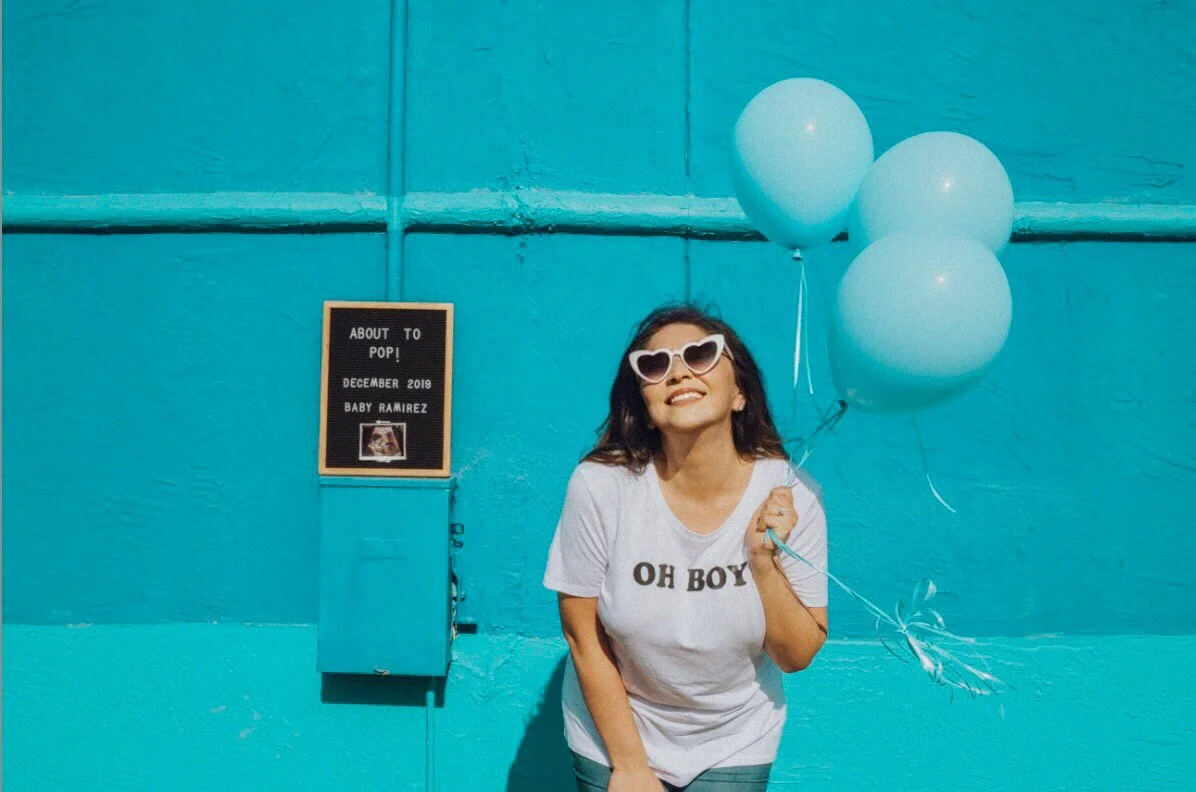 Woman with dark hair wearing heart-shaped sunglasses and a white T-shirt with 'OH BOY' on it, holding three white balloons in front of a bright blue wall. A small sign on the wall reads 'ABOUT TO POP! DECEMBER 2019 BABY RAMIREZ' with a picture of a s