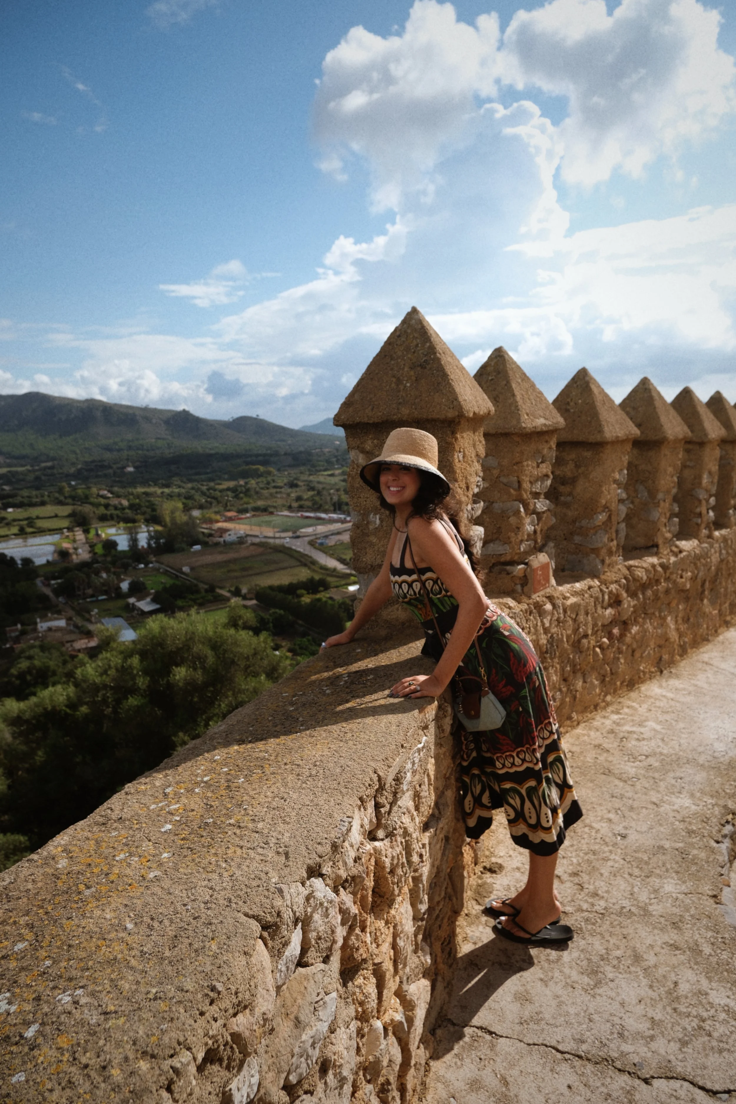 A woman in a sunhat and patterned dress is leaning on a stone wall with triangular crenellations, overlooking a scenic landscape with trees, rivers, and mountains under a partly cloudy sky.