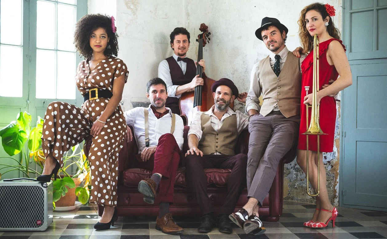Grupo de siete personas con ropa vintage, algunos con instrumentos musicales en un ambiente interior con paredes desgastadas y plantas.